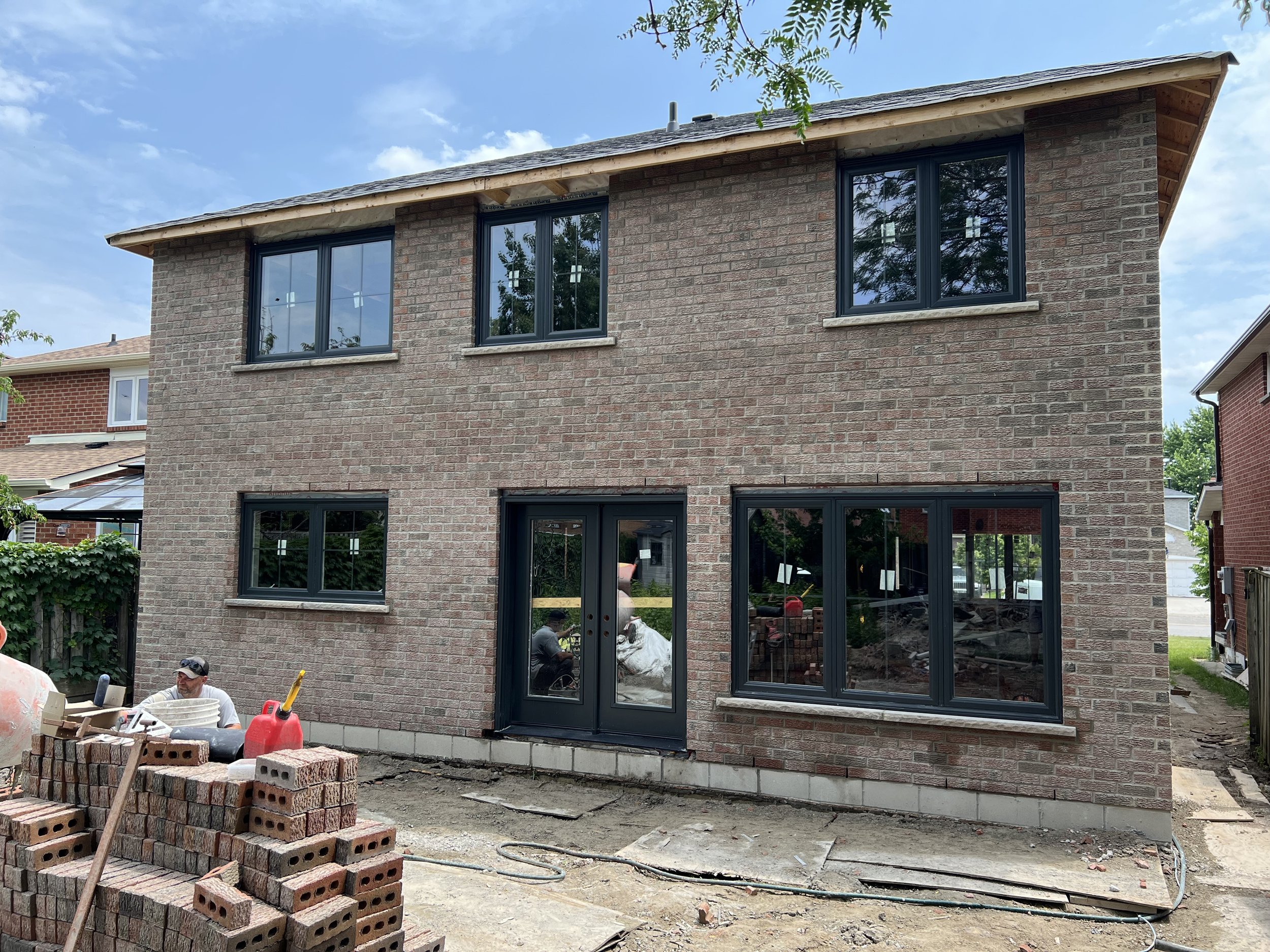 Two-story house under construction with brick exterior, multiple windows, and a partially installed door. Building materials and bricks are scattered in the foreground. A construction worker is present near a cement mixer and a stack of bricks. The s