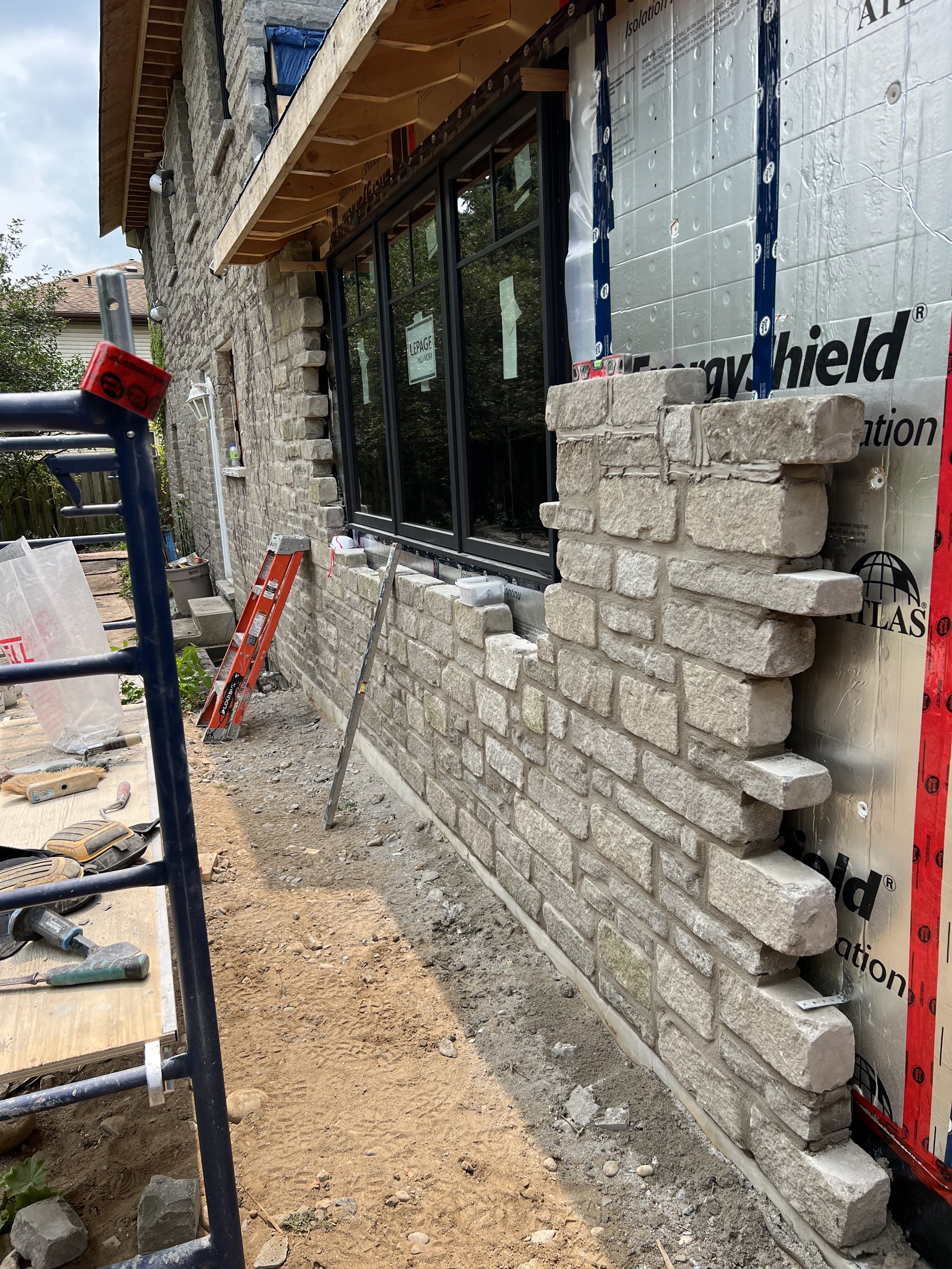 Construction site with partially built stone wall on a building, featuring a window. Tools and scaffolding are visible, along with insulation materials.