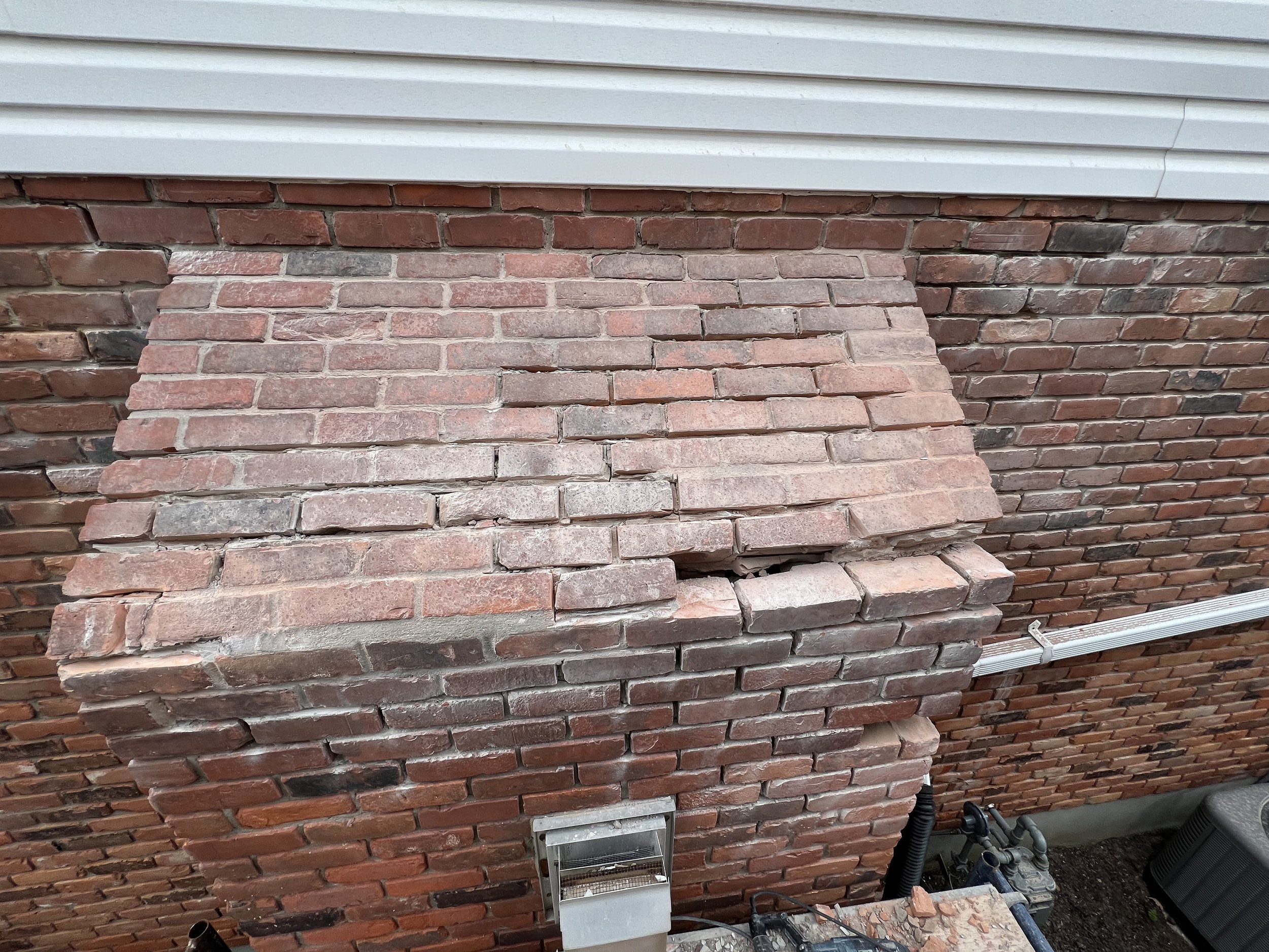 A damaged brick chimney with visible cracks and misaligned bricks attached to a brick house exterior.
