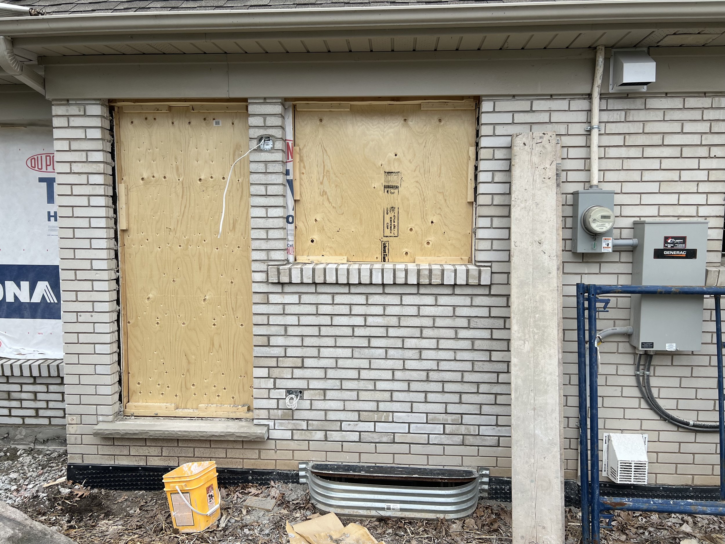 Brick wall with boarded-up door and window, yellow bucket on the ground, electrical meter box, and wooden plank leaning against the wall.