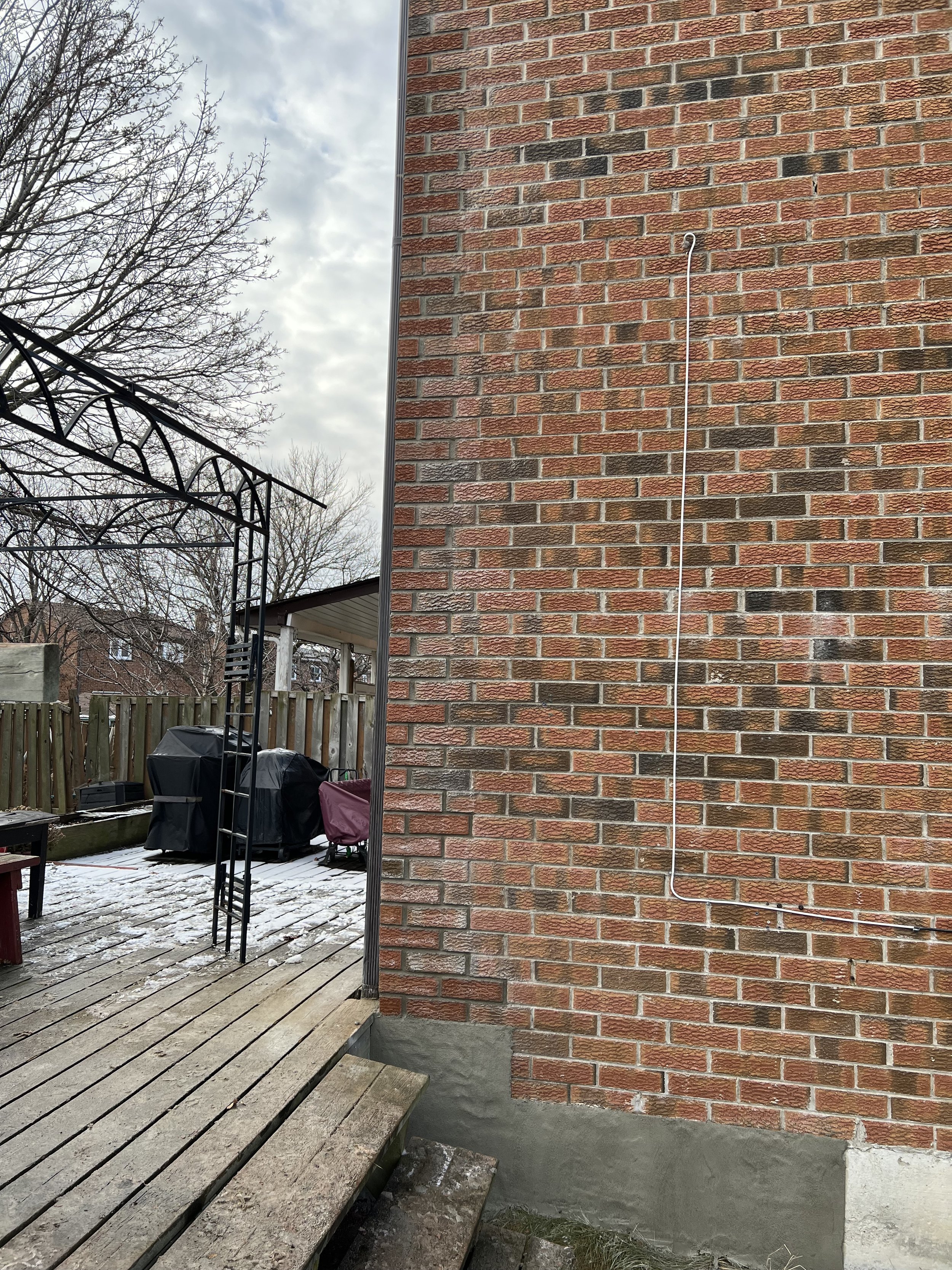 Exterior red brick wall with a metal wire and nearby deck, snow on the ground, bare trees, and outdoor furniture.