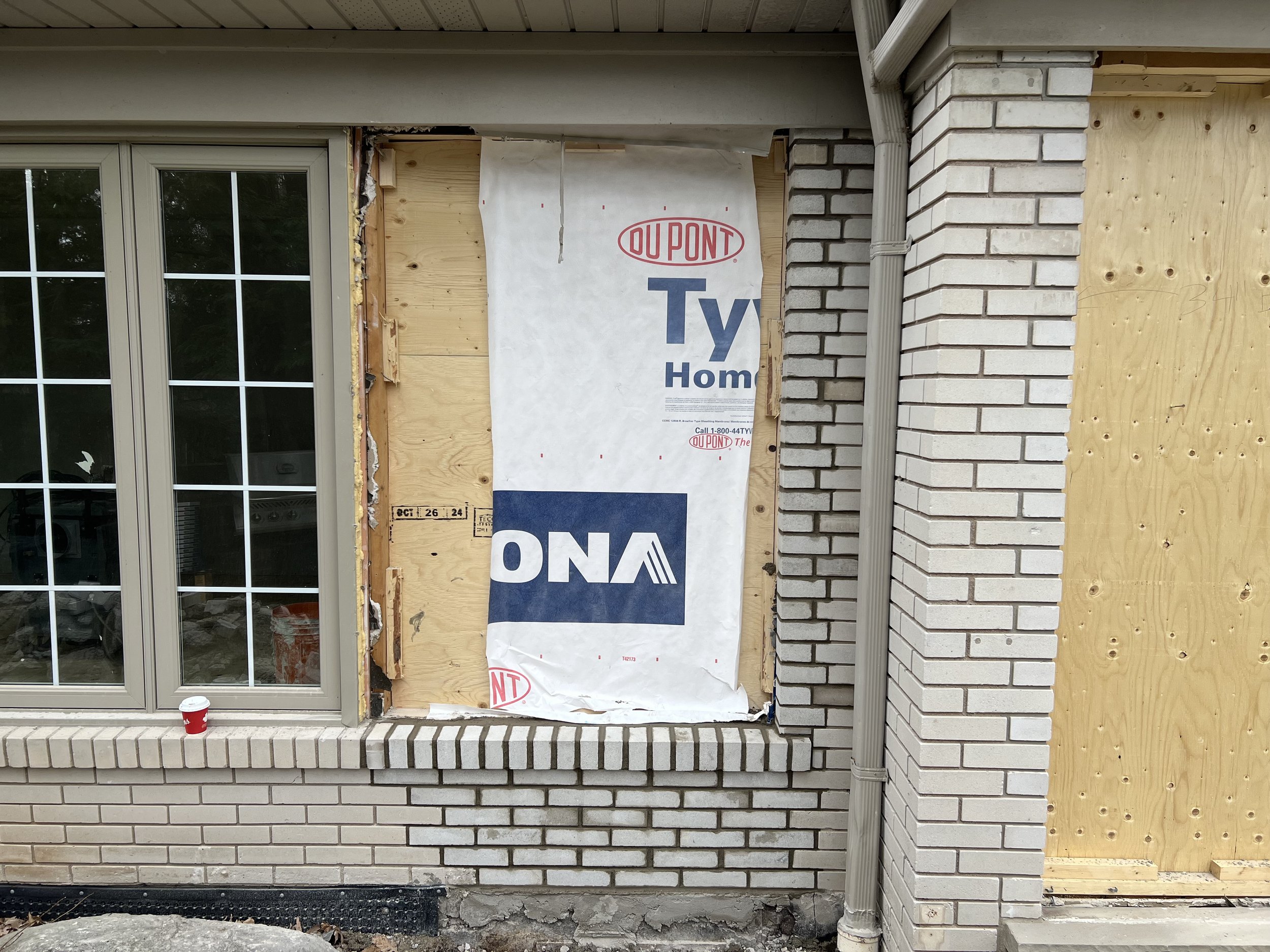 House undergoing exterior renovation with plywood and Tyvek wrap, adjacent to a window and brick wall; a red cup sits on the brick ledge.
