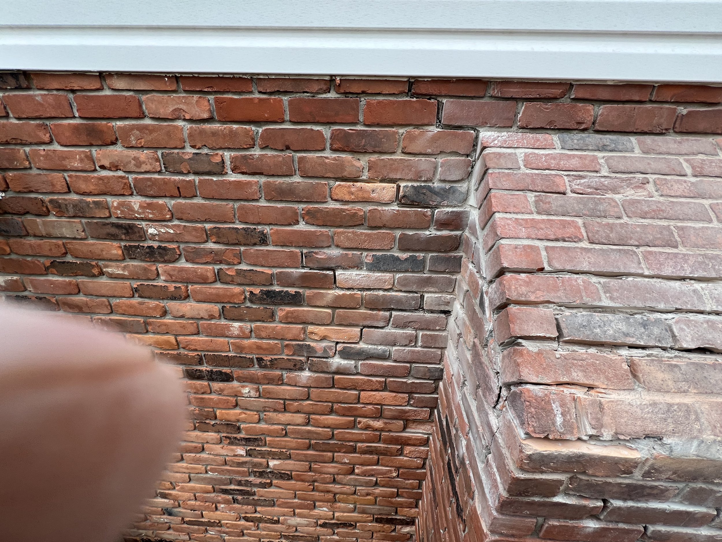 Brick wall with visible cracks and discolored areas, featuring a protruding section and part of a finger in the lower left corner.