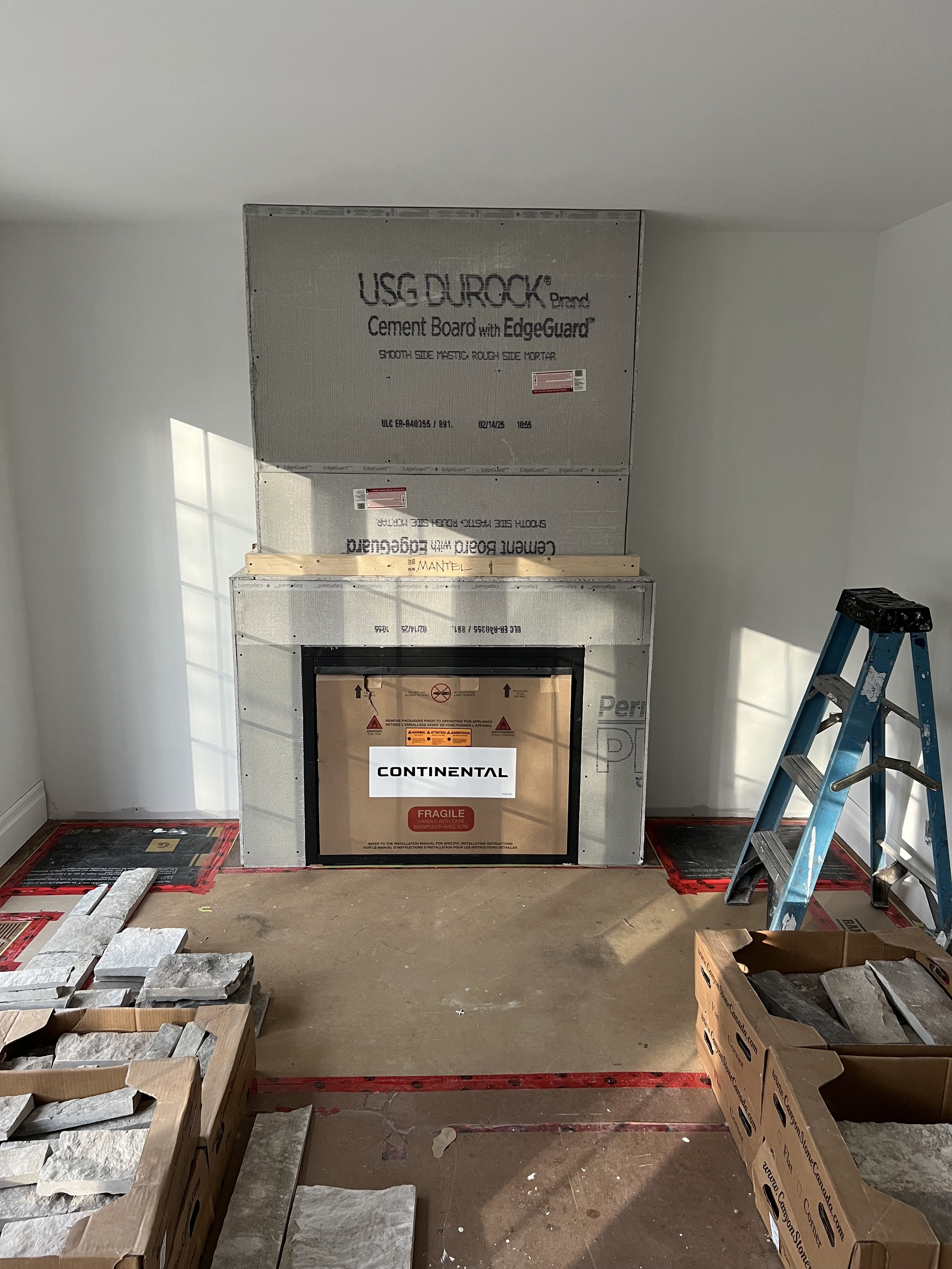Incomplete fireplace installation with cement board, blue ladder, and stone veneer boxes on the floor.