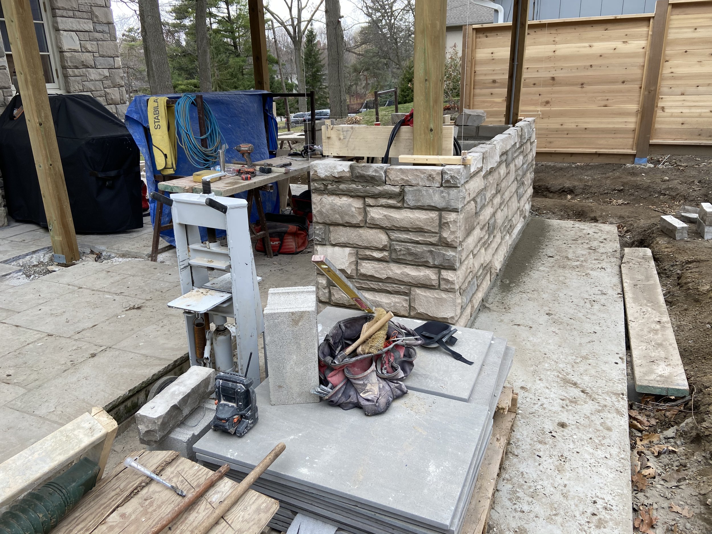 Construction site with stacked stone wall, workbench, tools, and wooden structure in a yard.