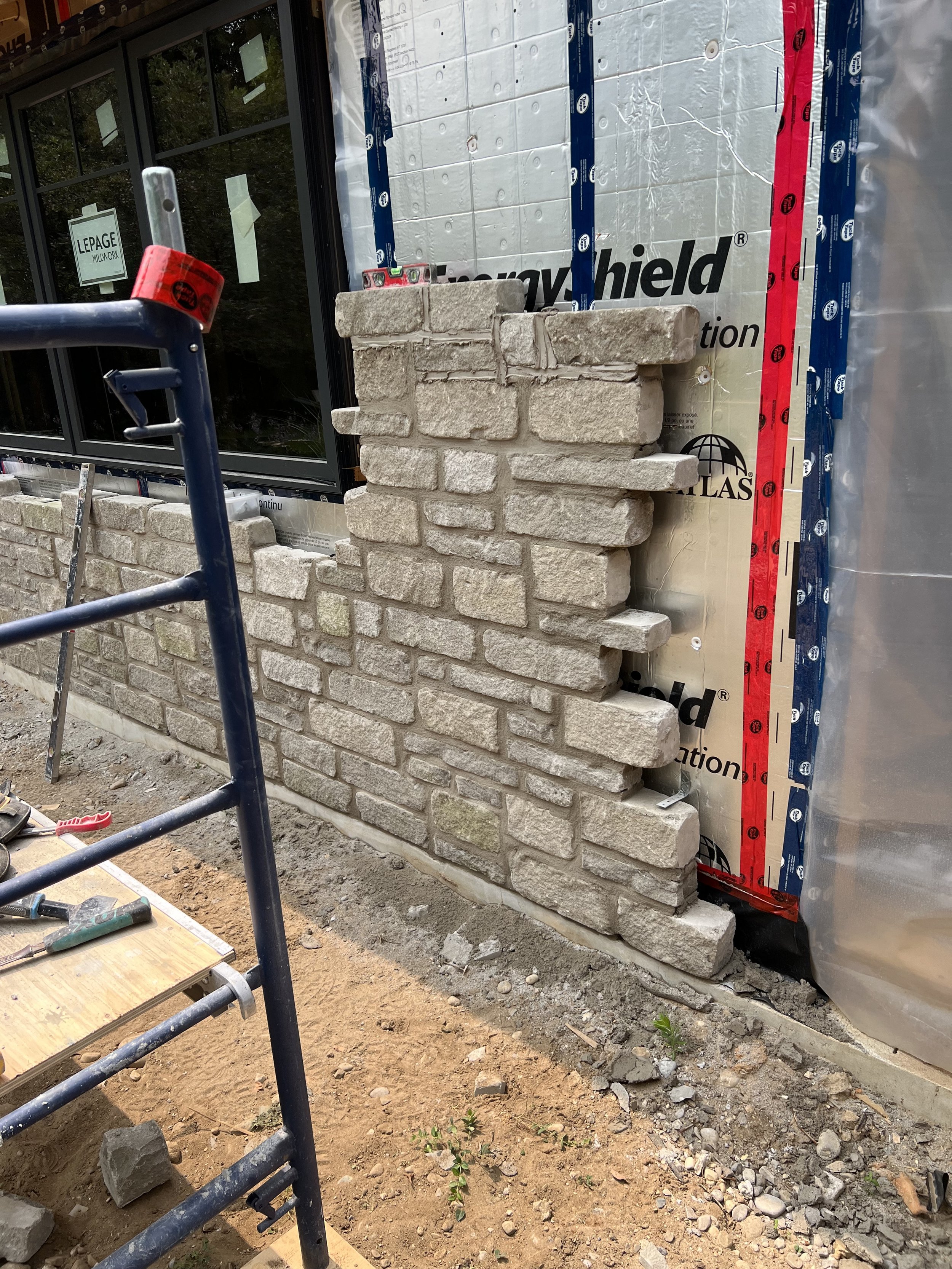 Partial stone wall under construction with exposed insulation and partially built brickwork. A scaffold is adjacent to the wall. The ground is covered with dirt and stones.
