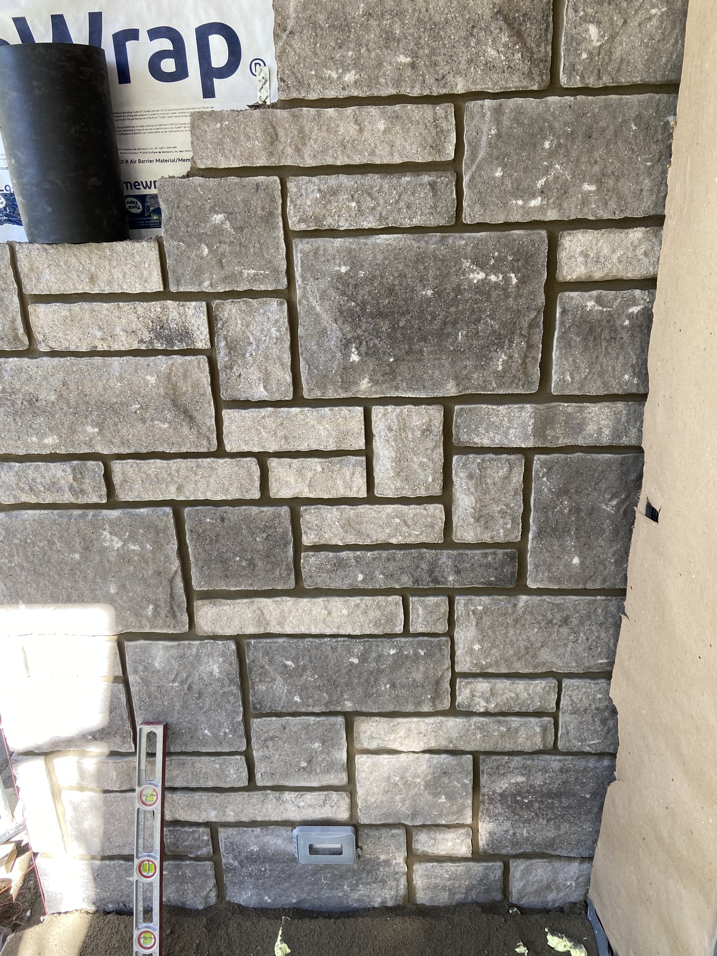 Stone veneer wall with staggered rectangular stones, a level tool leaning against it, and a partially visible paper wrap on top.