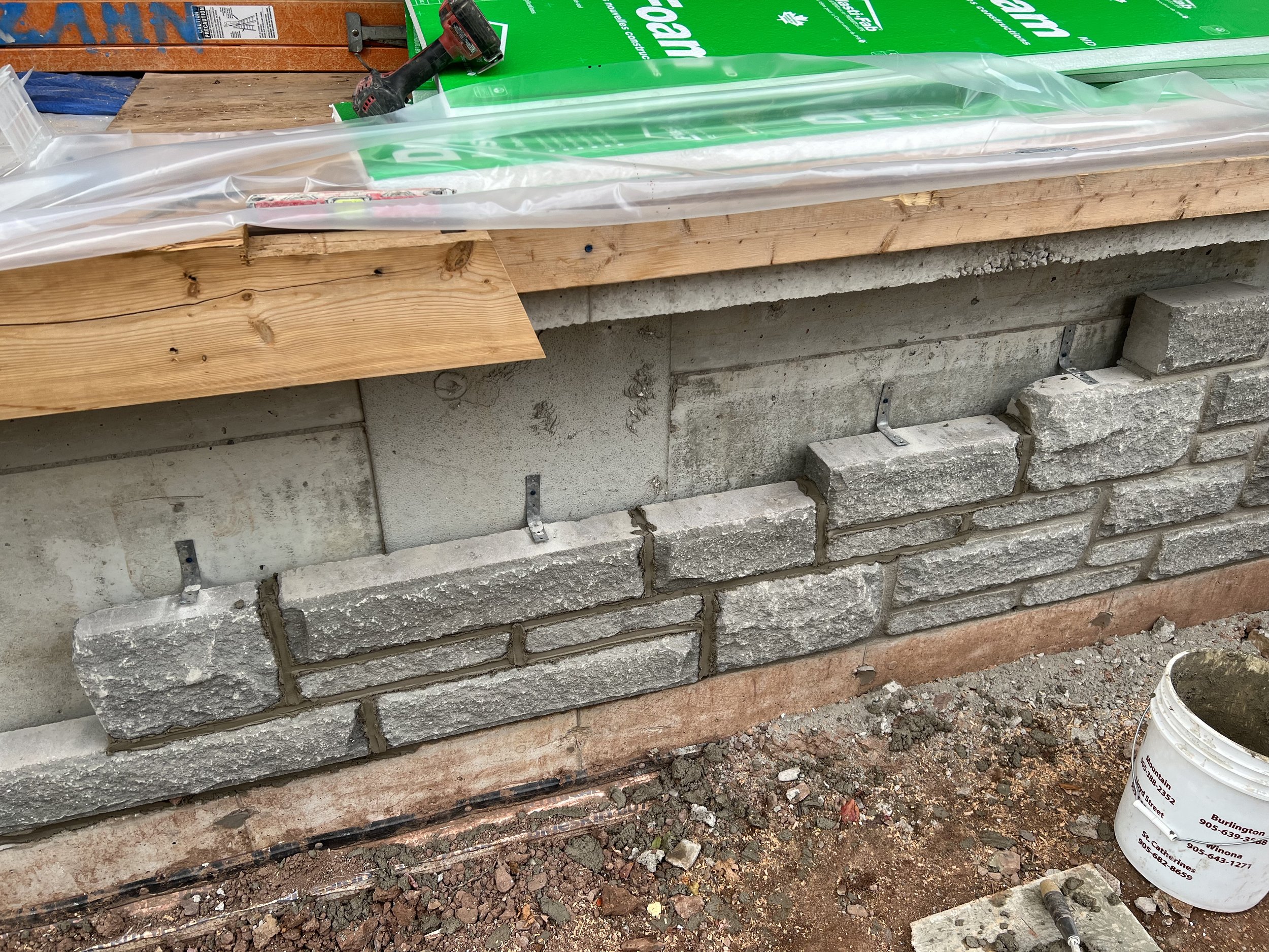 Construction of a stone veneer wall with metal brackets, wooden boards, insulation materials, and tools like a drill and bucket nearby.