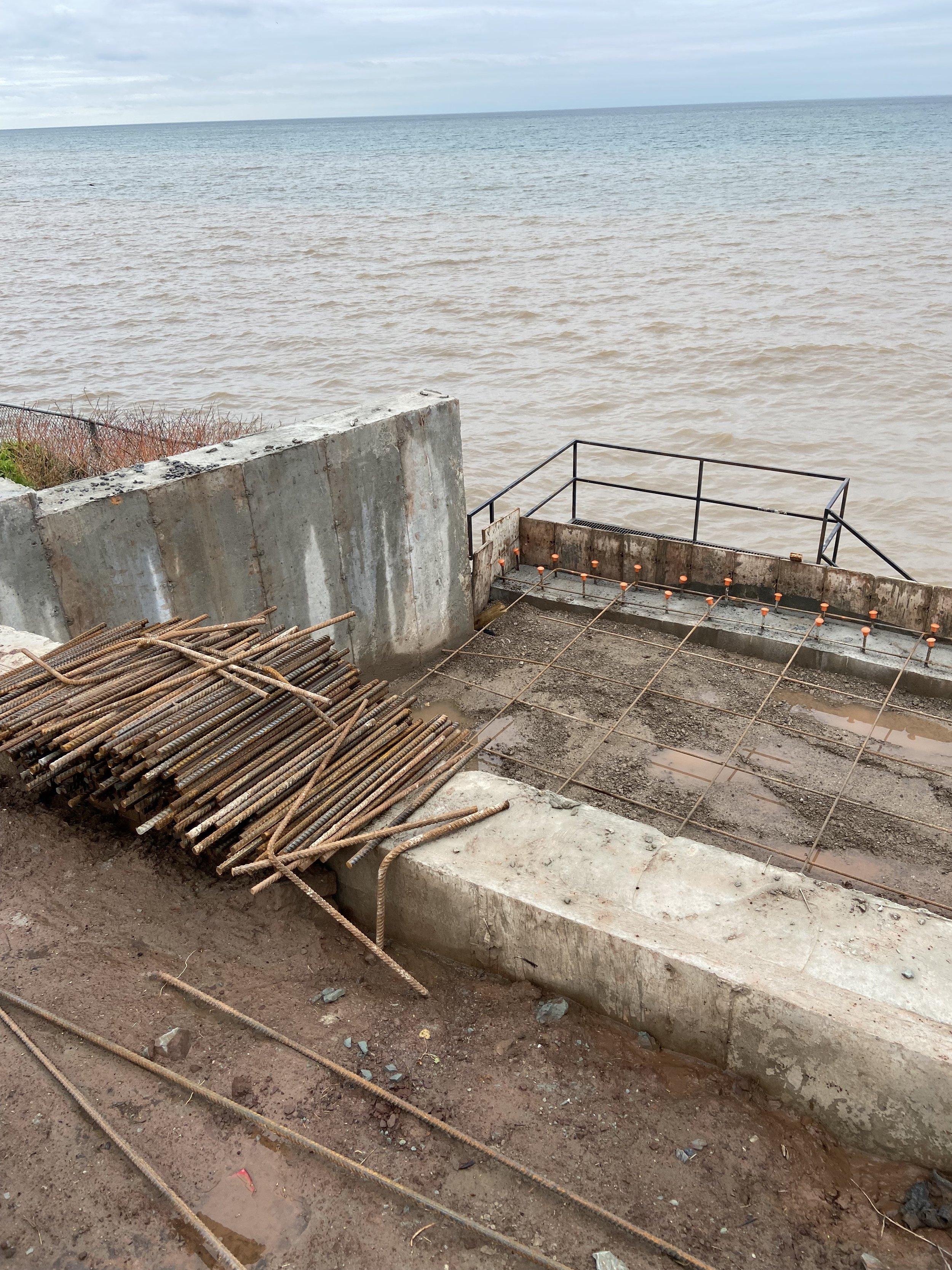 Construction site by a body of water with rebar rods and concrete structures.