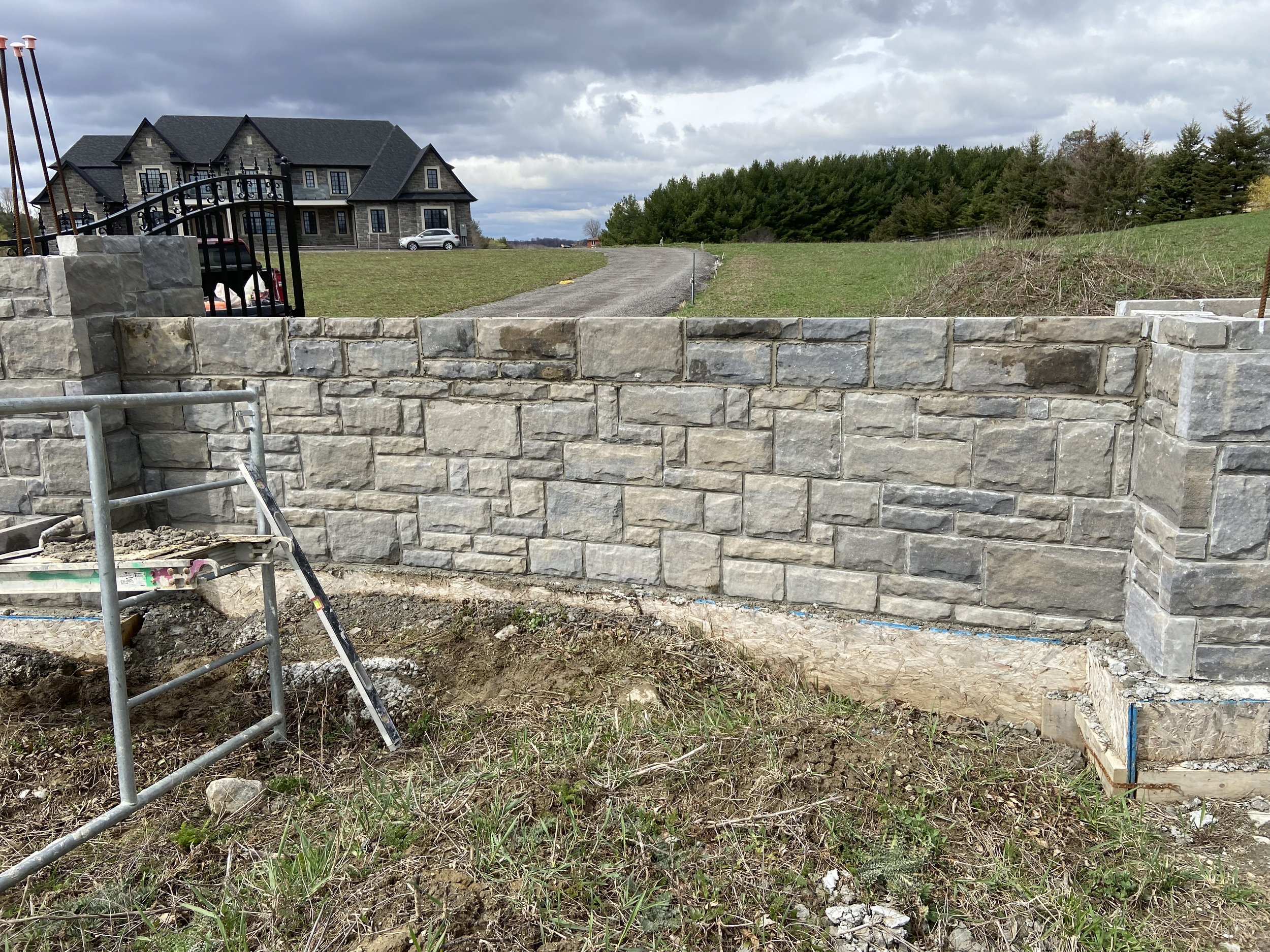 Stone wall under construction with scaffolding in front, grass and dirt in the foreground, large house and driveway in the background, cloudy sky.