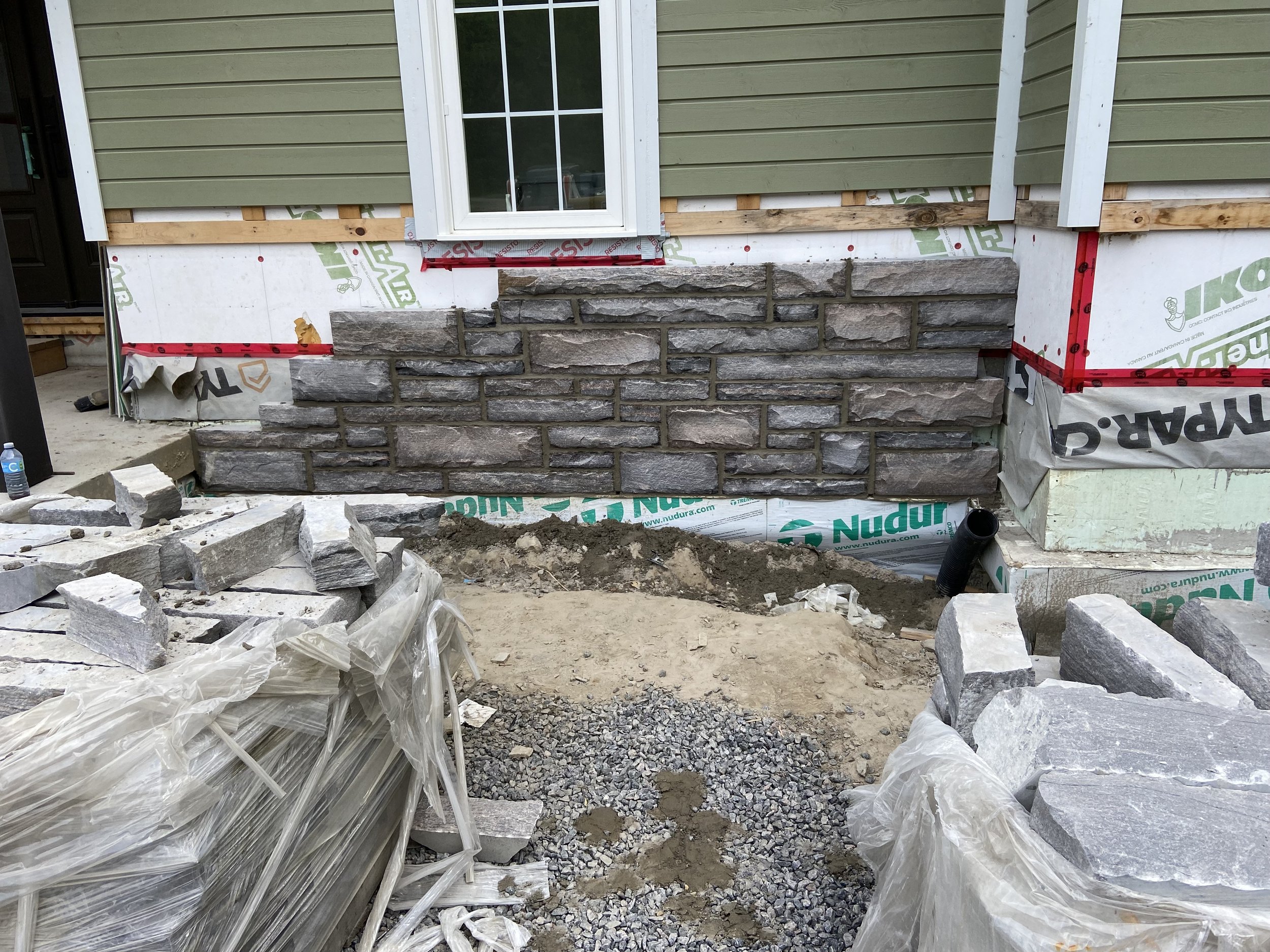 House exterior under construction with partially finished stonework, siding, and wrapping materials.