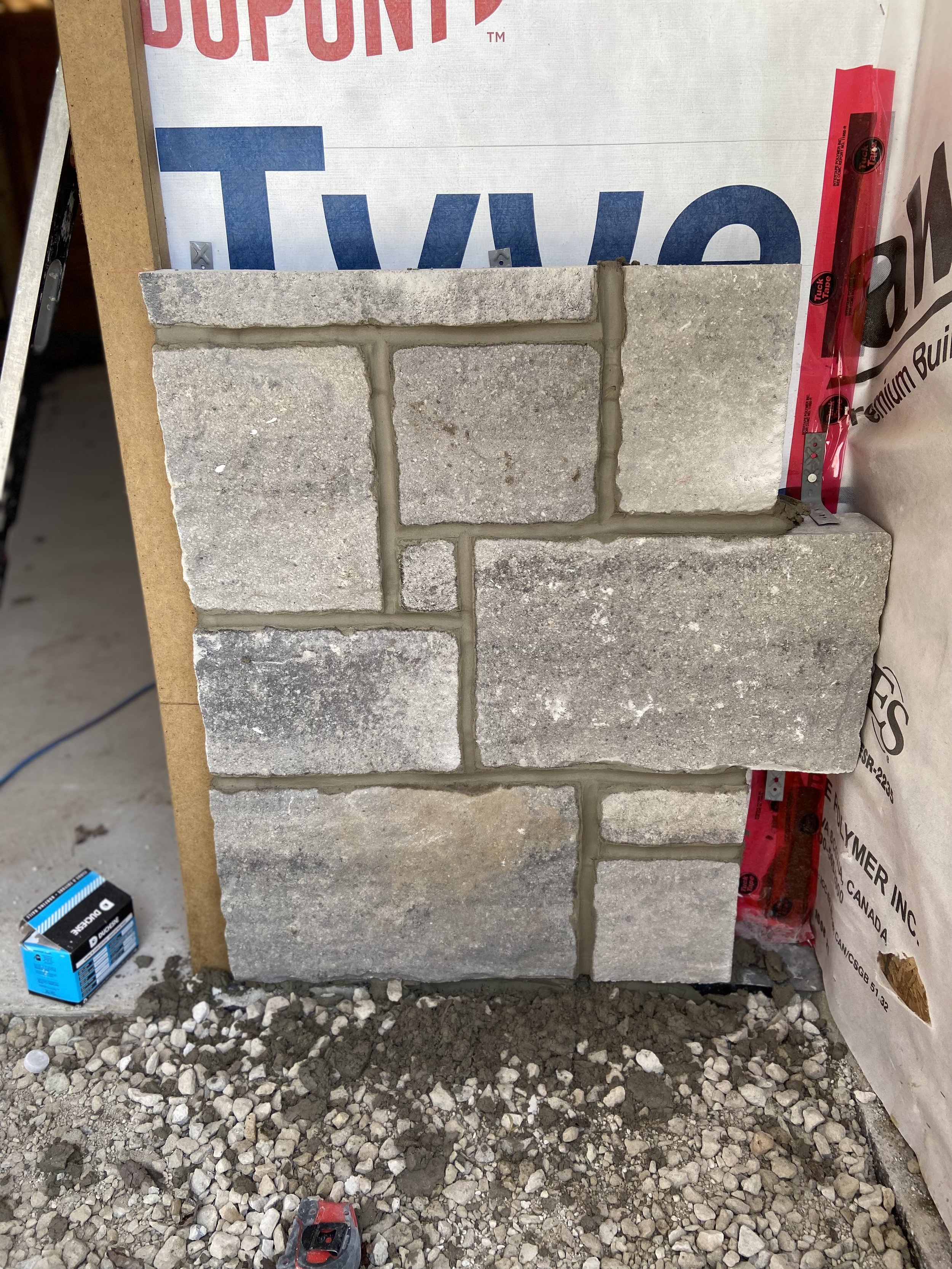 Stacked stone veneer installation in progress against a wall with Tyvek and other building materials visible.