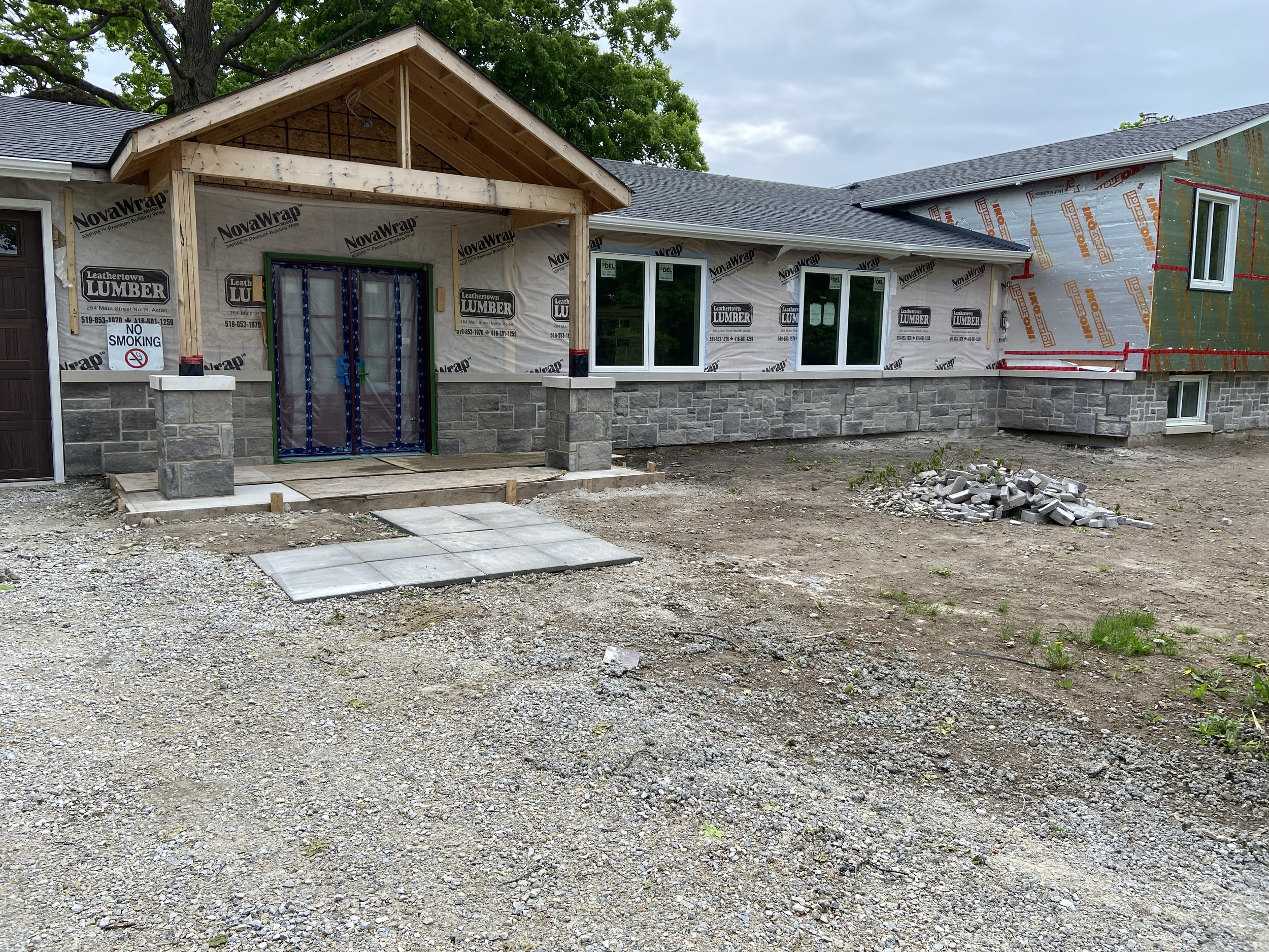 House under construction with stone facade and unfinished entrance, covered with building wrap material, surrounded by a gravel yard.