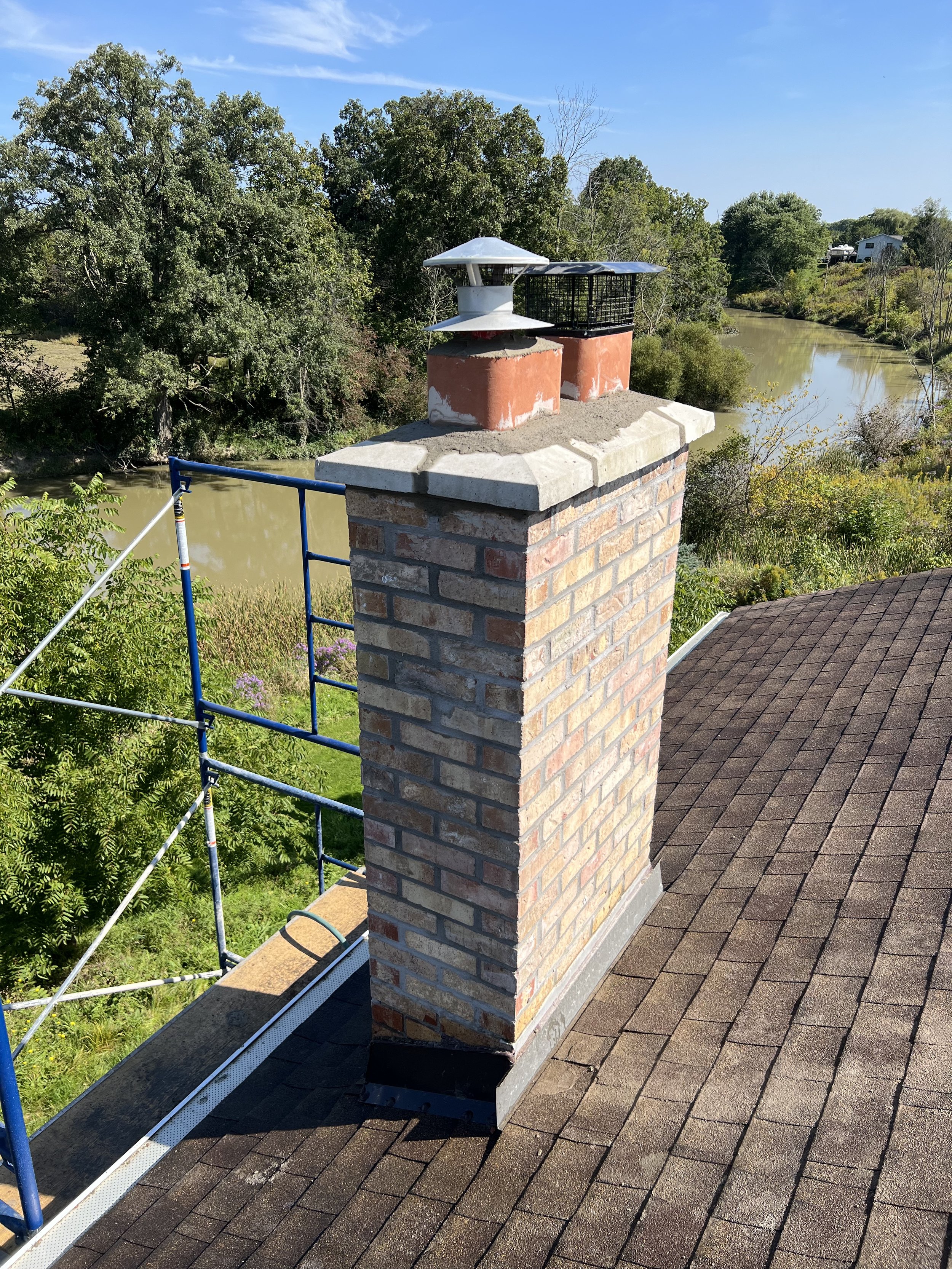 A brick chimney with two metal chimney caps on a roof, next to a scaffold. Trees and a river in the background.