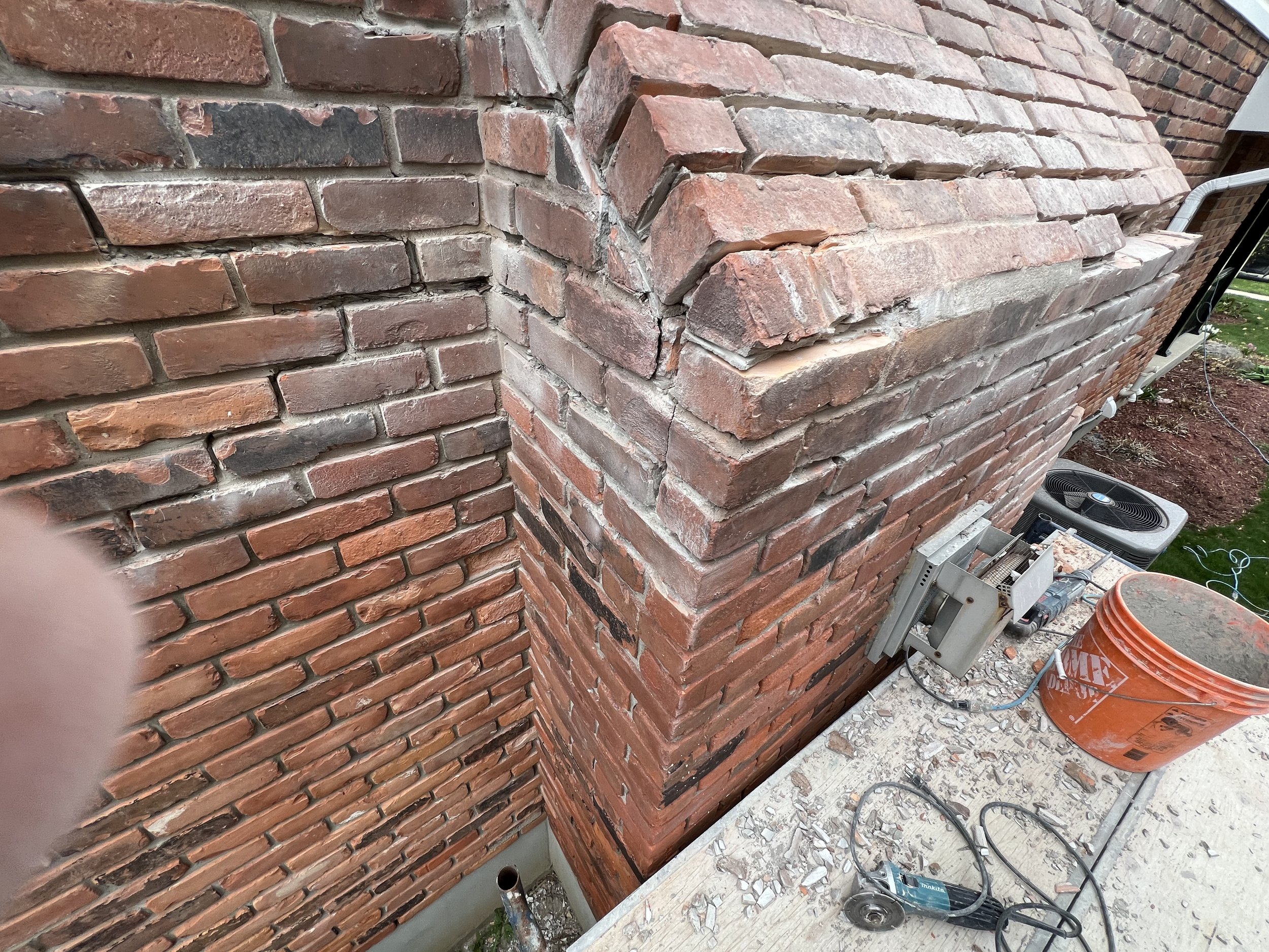 Damaged brick chimney with cracks and missing mortar, surrounded by construction tools and debris on a wooden platform. An HVAC unit and plastic bucket are visible nearby.