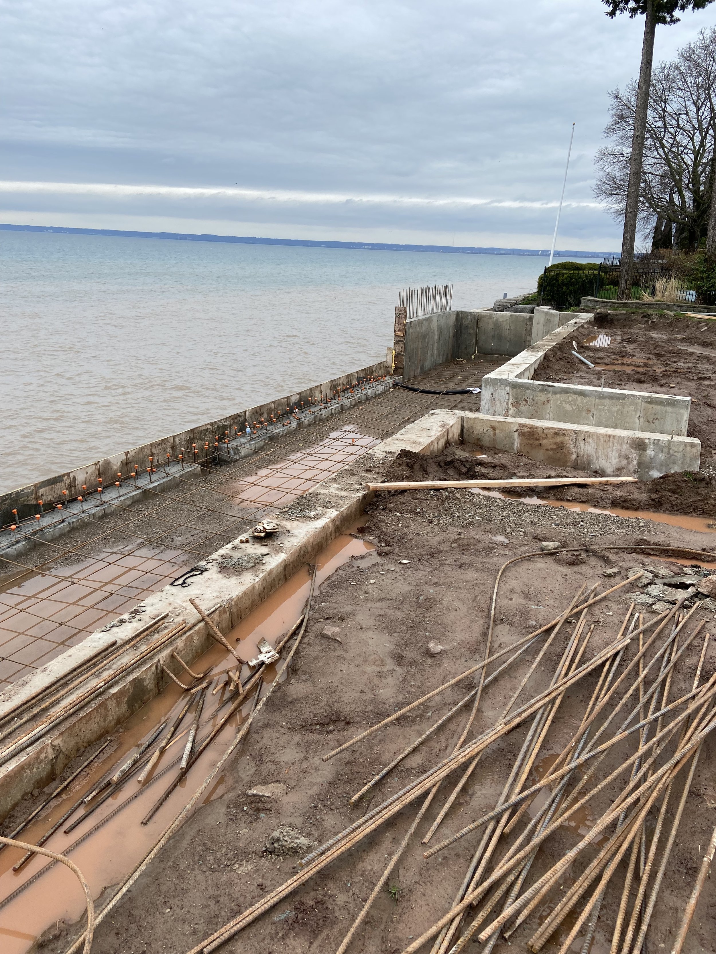 Seawall construction site by a lake with rebar and concrete forms, overcast sky, and trees on the side.