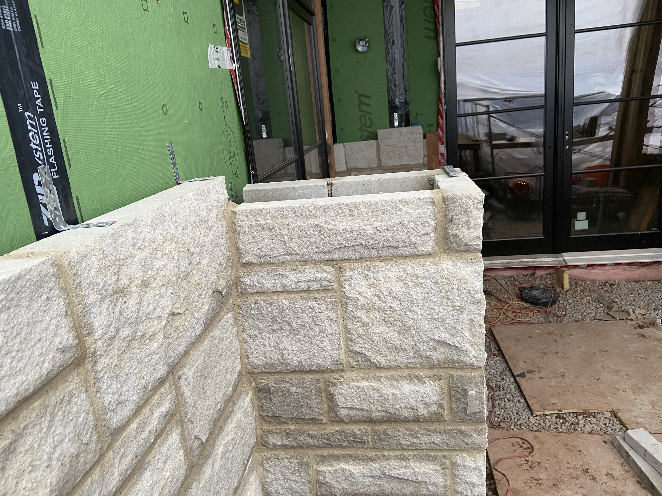 Close-up of a stone wall under construction with green sheathing and black framing tape, adjacent to a glass door.