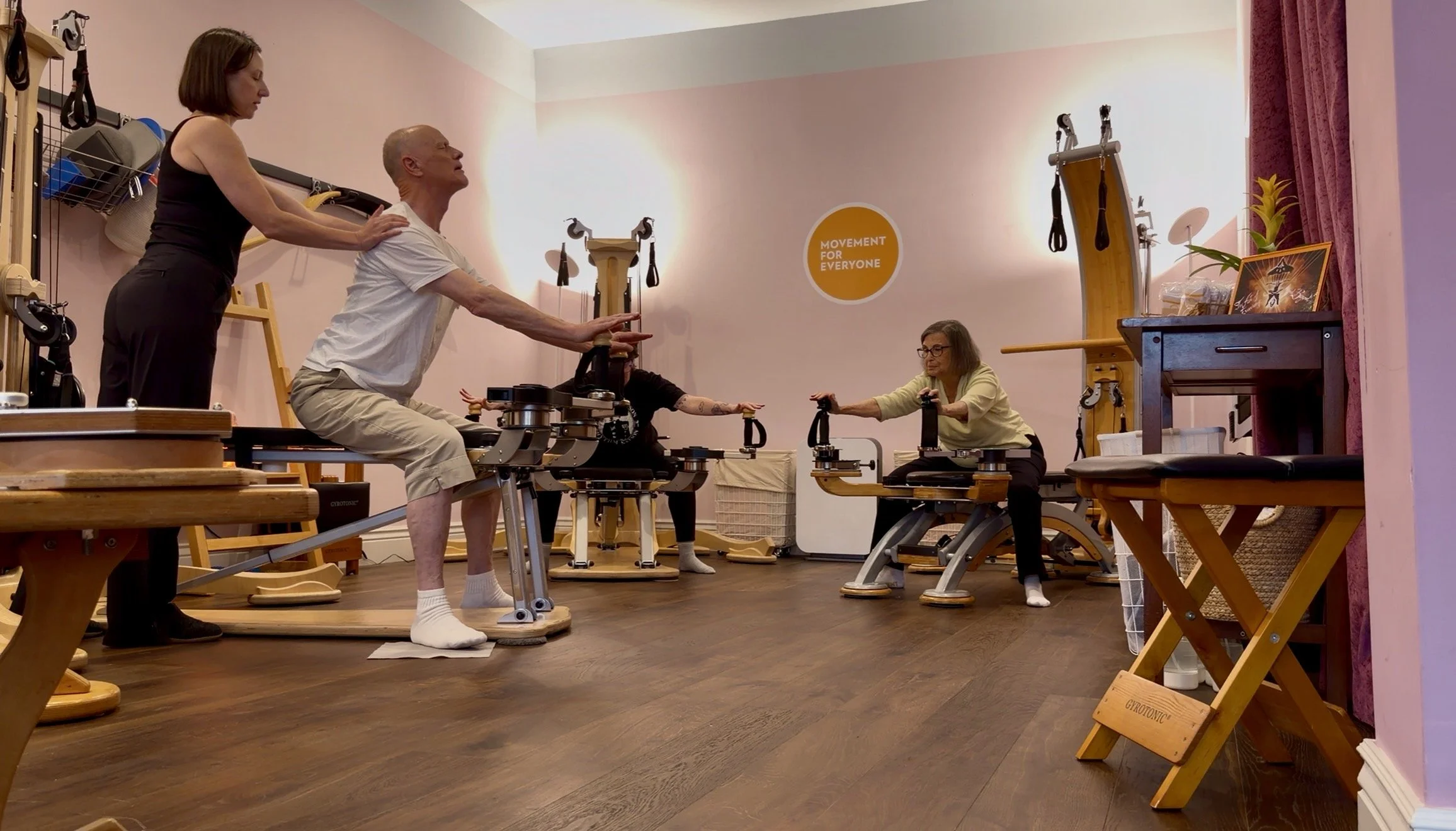 Three people practicing Gyrotonic exercise at a studio, seated with arms extended forward and hands holding the Gyrotonic handles. An instructor is providing hands-on cuing to a man's shoulders.