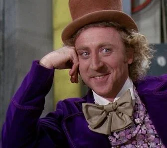 Image of Gene Wilder as Willy Wonka wearing his chestnut brown top hat, royal purple coat, and beige bowtie nearly ready to fly away. He is resting his hand on his temple with an impish grin on his face and a twinkle in his eye.
