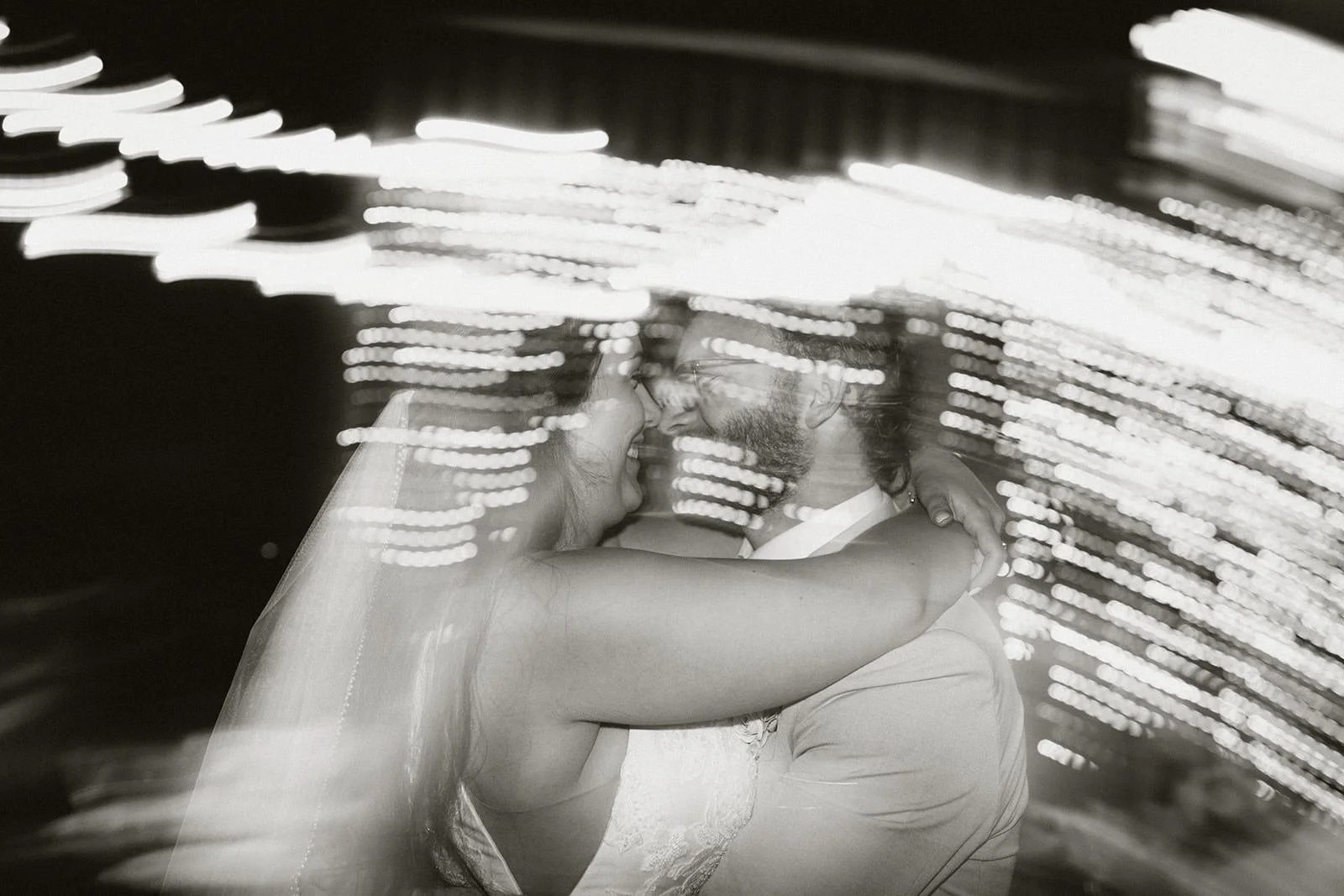 A couple hugging and smiling at each other, illuminated with blurred bright lights in the background.