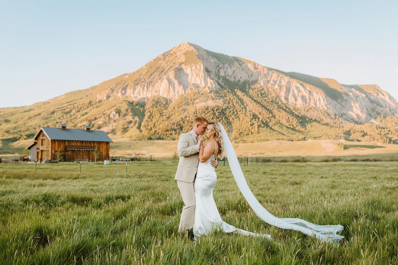 A newlywed couple celebrating by a lake with mountains in the background. The groom is running and spraying water from a bottle, while the bride, in a white lace dress, is holding a bouquet and celebrating with arms raised.