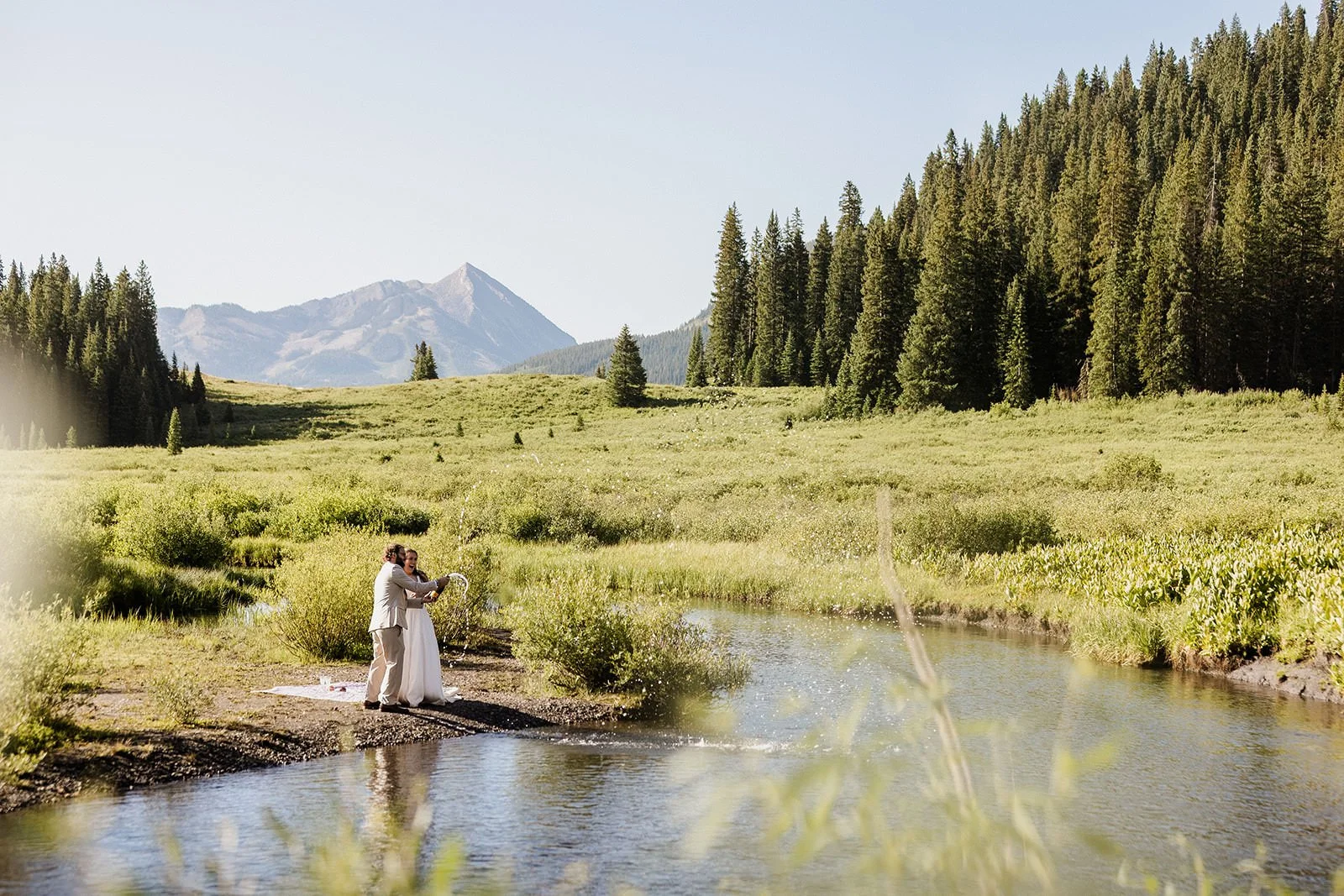A couple getting married in a scenic outdoor setting with mountains, forests, and a small river.