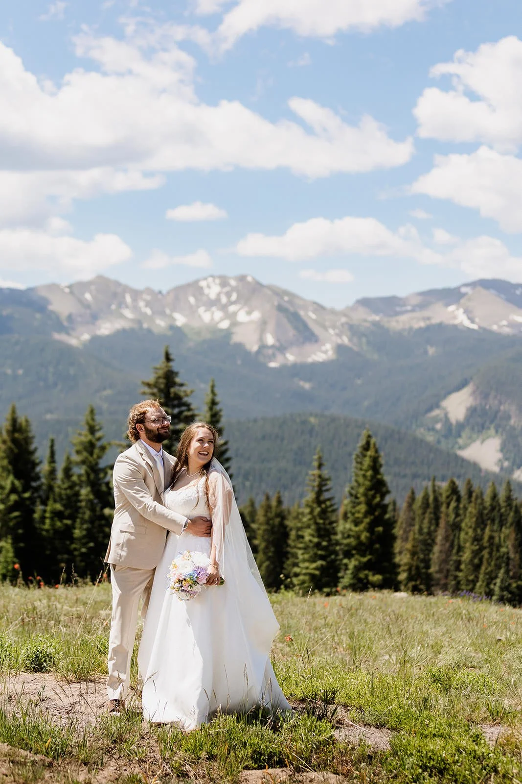 A bride and groom standing in a field with mountain and forest backdrop, the groom with his arm around the bride who is holding a bouquet, both smiling and looking happy.