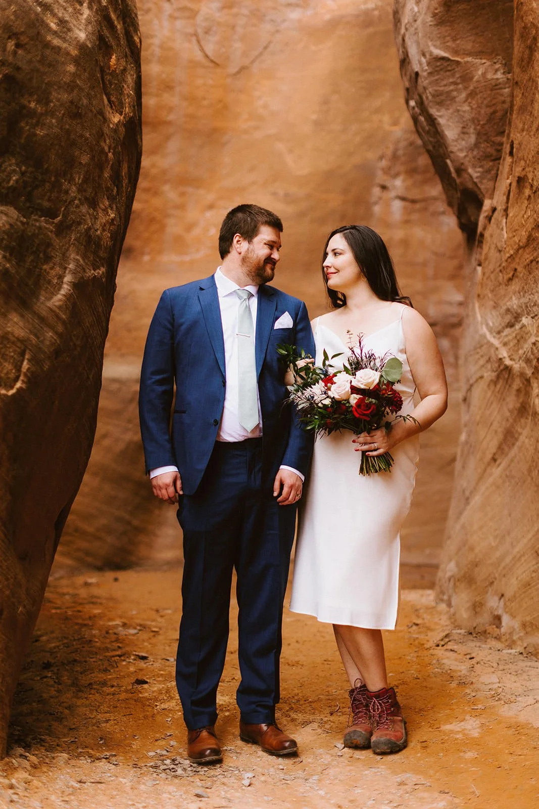 Short white elopement dress worn with hiking boots, next to a navy suit in a red rock slot canyon