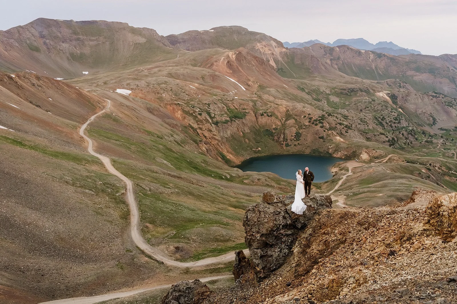 A bride and groom standing on a rocky ledge in a mountainous landscape with winding trails, green patches, and a lake in the background.