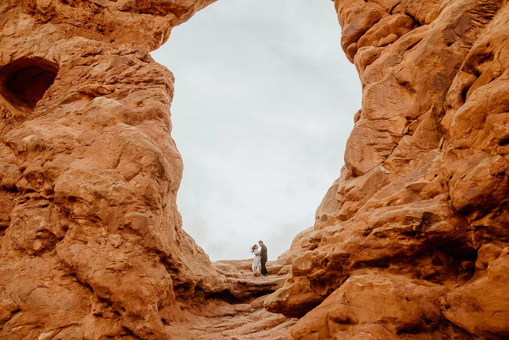 A couple stands under a huge red sandstone arch in Moab
