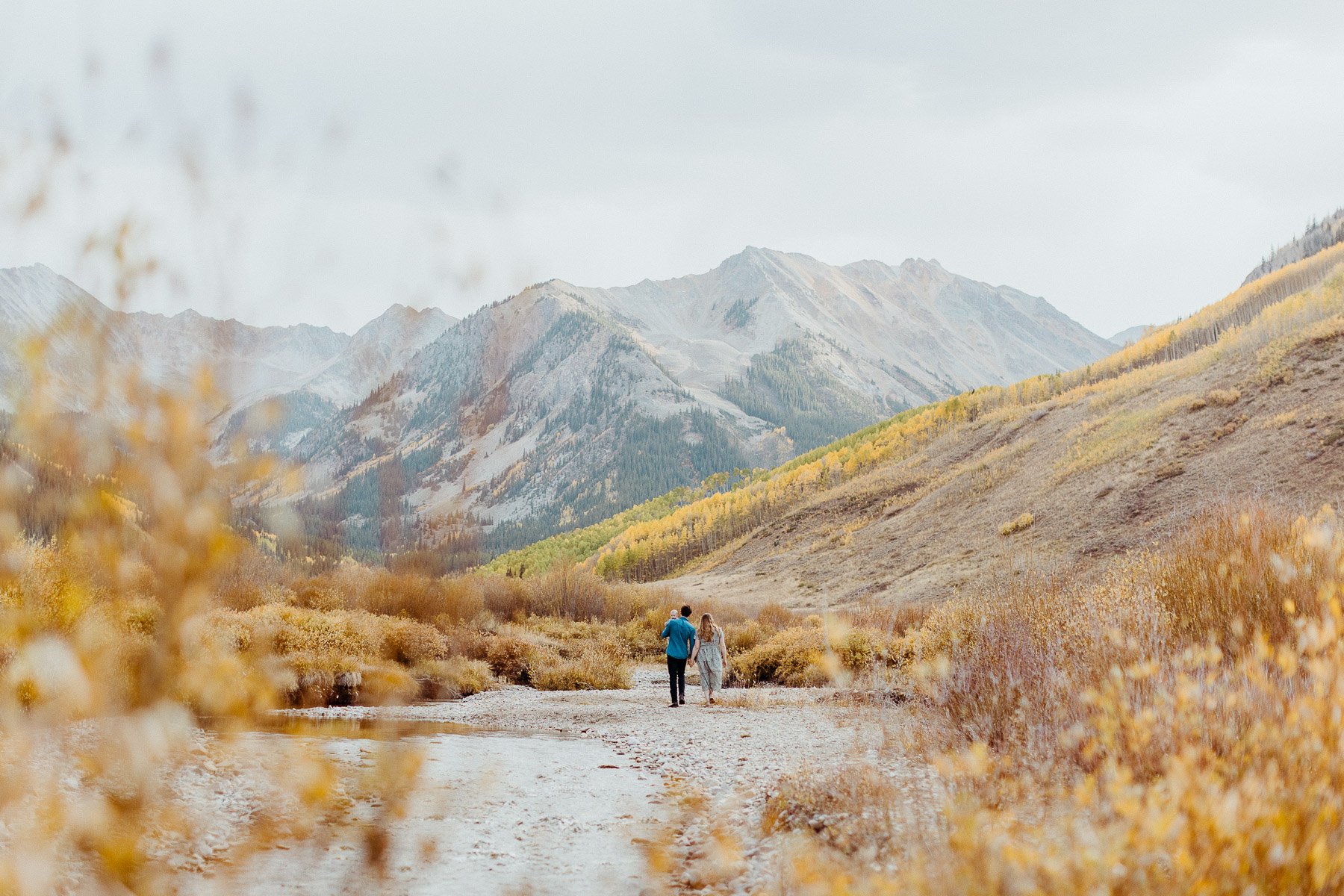 A couple walking hand in hand on a dirt trail through a mountain valley with autumn foliage and rugged peaks in the background.