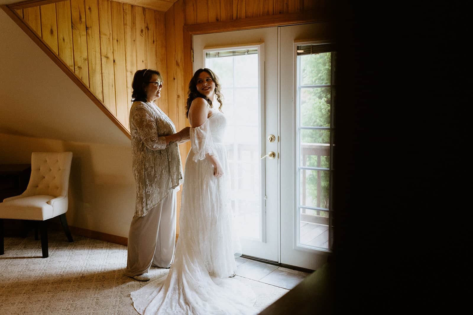 A mother helps her daughter put on her wedding dress in front of a window with natural light