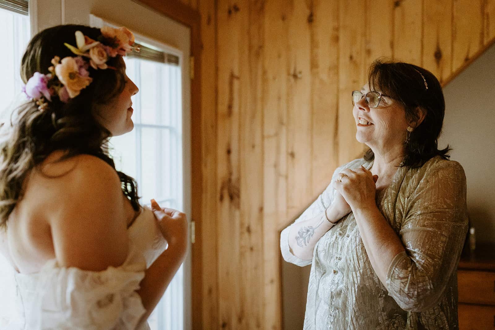 A mother holds her hands tight in a clap and smiles at her daughter as she sees her in her wedding dress for the first time