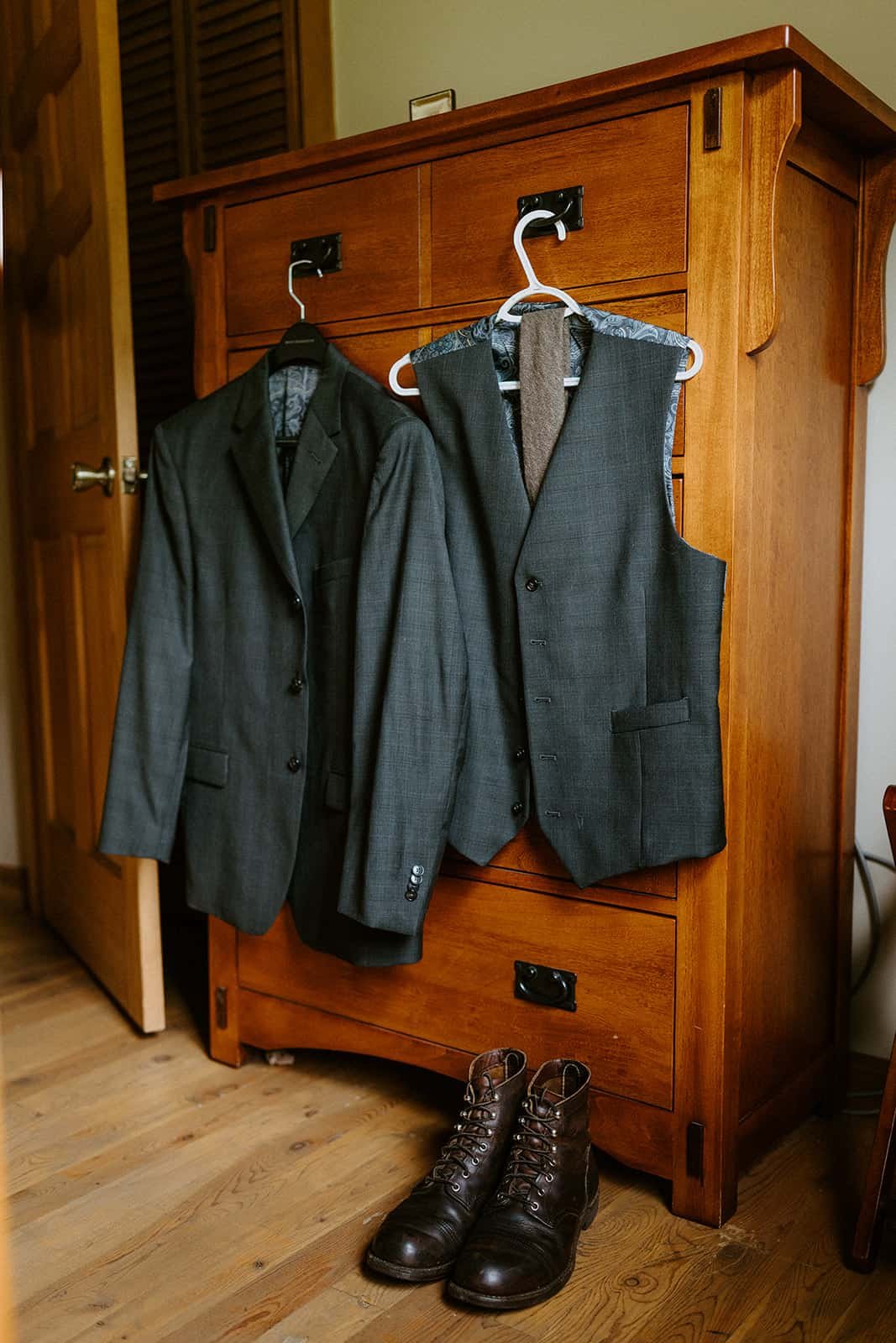 A suit and jacket hang on a wood dresser in front of Redwing boots