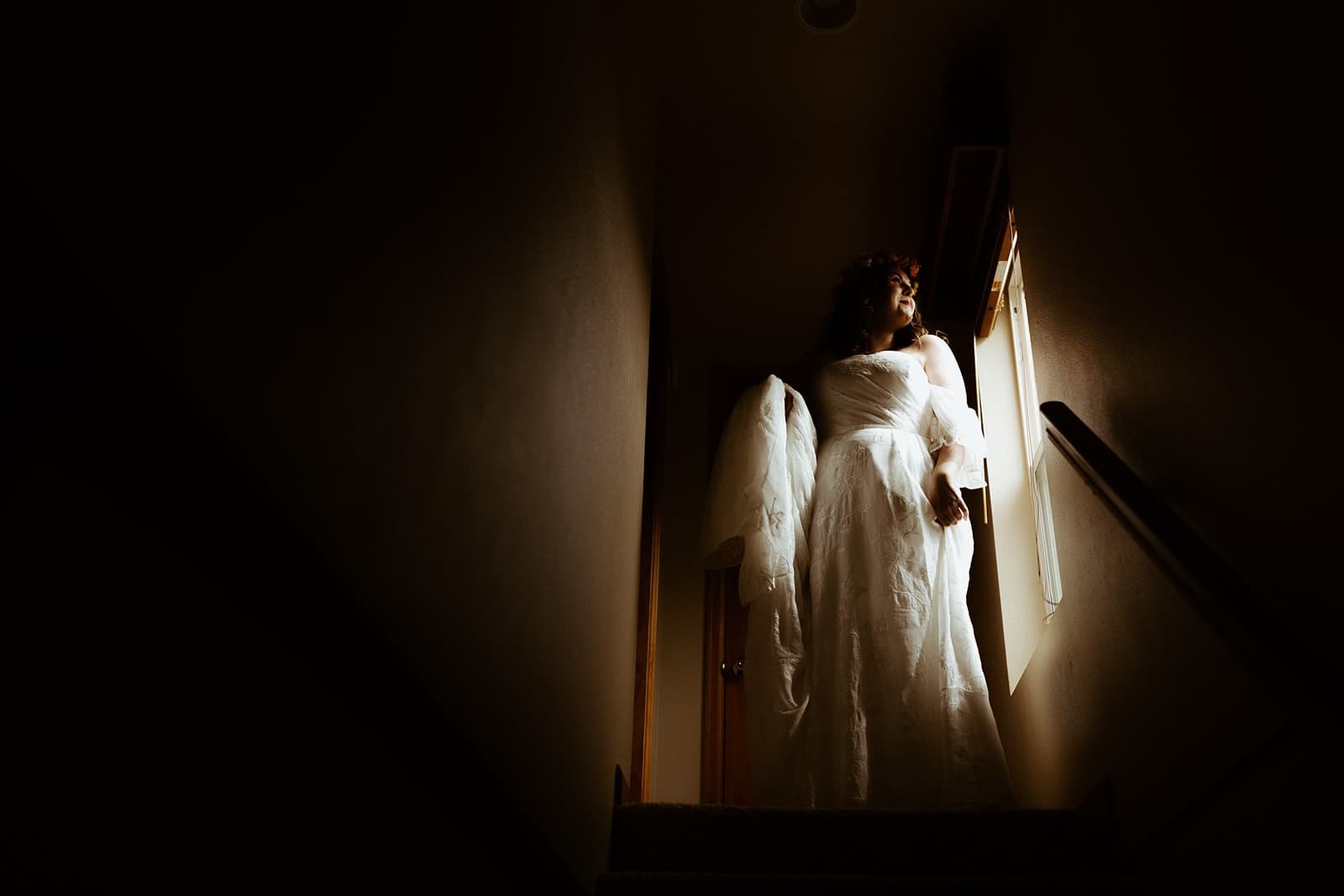 A bride descends a staircase as a window shines natural light on her in her wedding dress