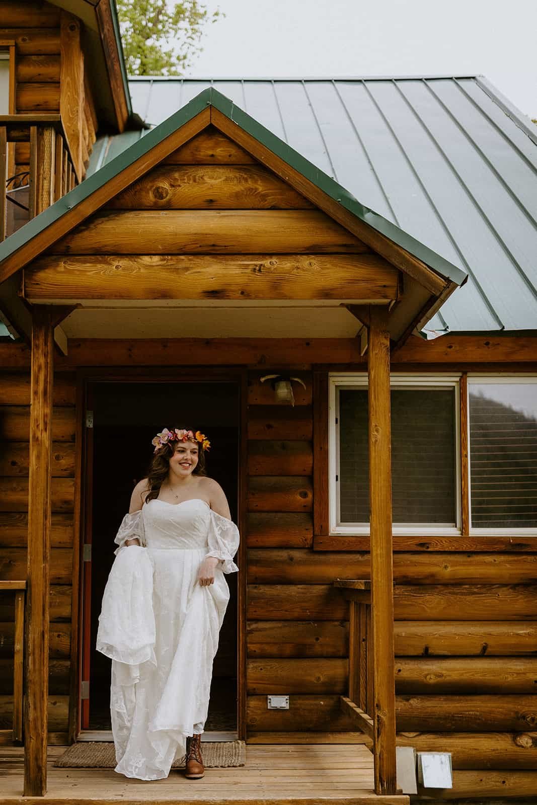 A woman holds her wedding dress up as she walks outside of the front door of a cabin