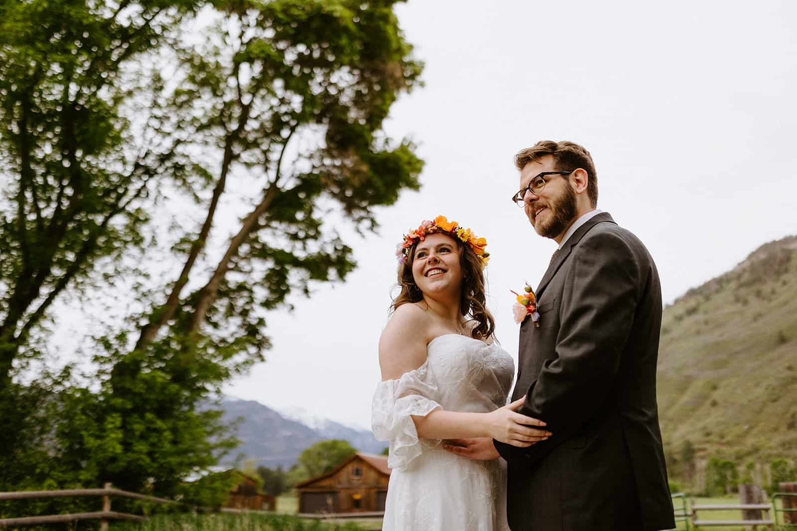 A man and woman look to the left smiling in front of a barn and mountains