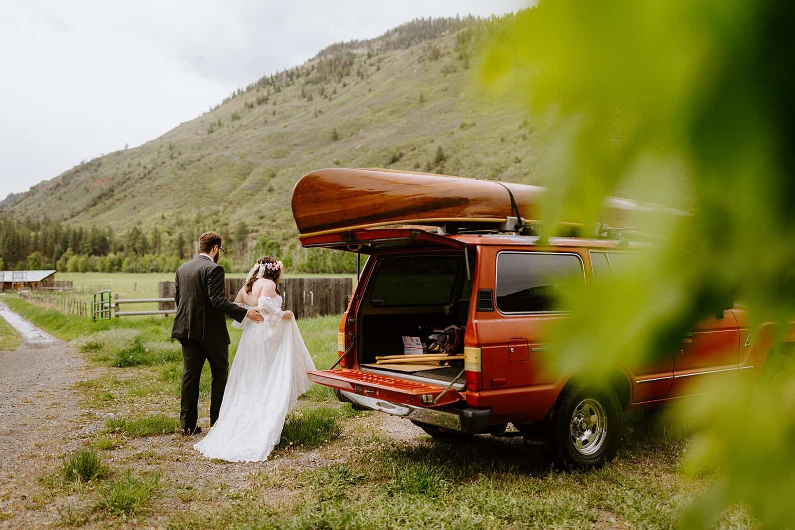 A man and woman walk around a red Toyota Land Cruiser with a wood canoe strapped to the roof in their wedding clothes