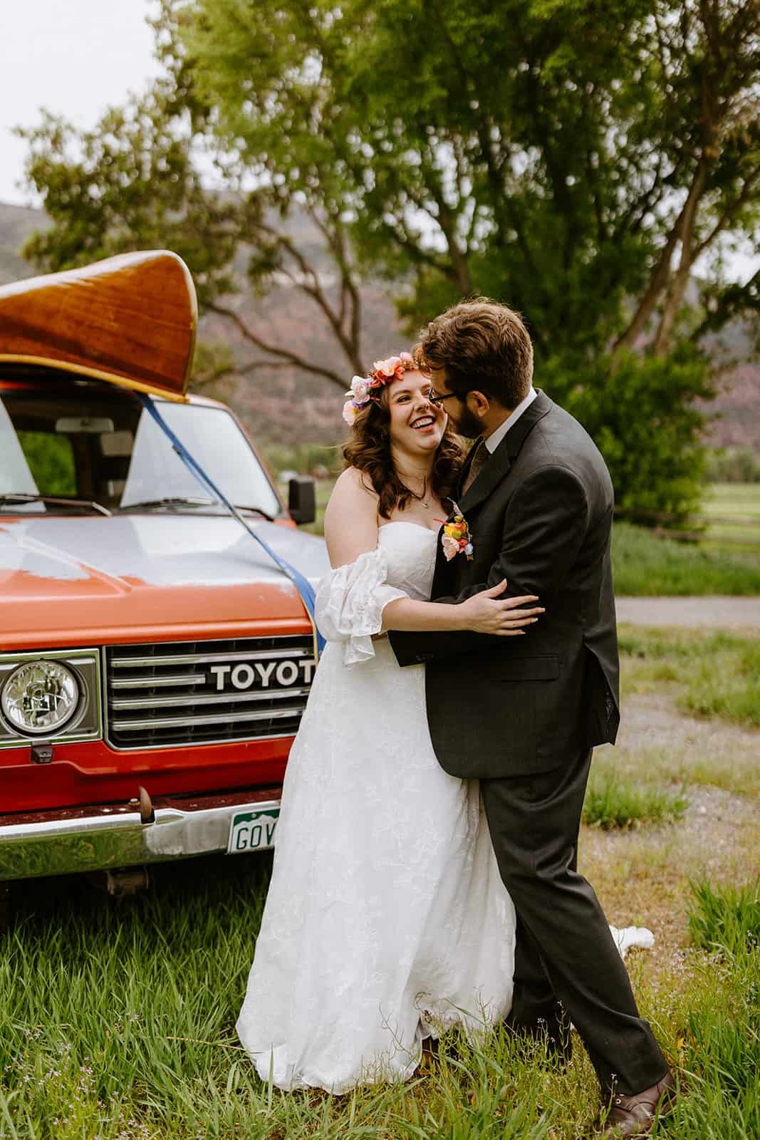 A man and woman in a wedding dress and suit smile and embrace in front of a red Toyota Land Cruiser