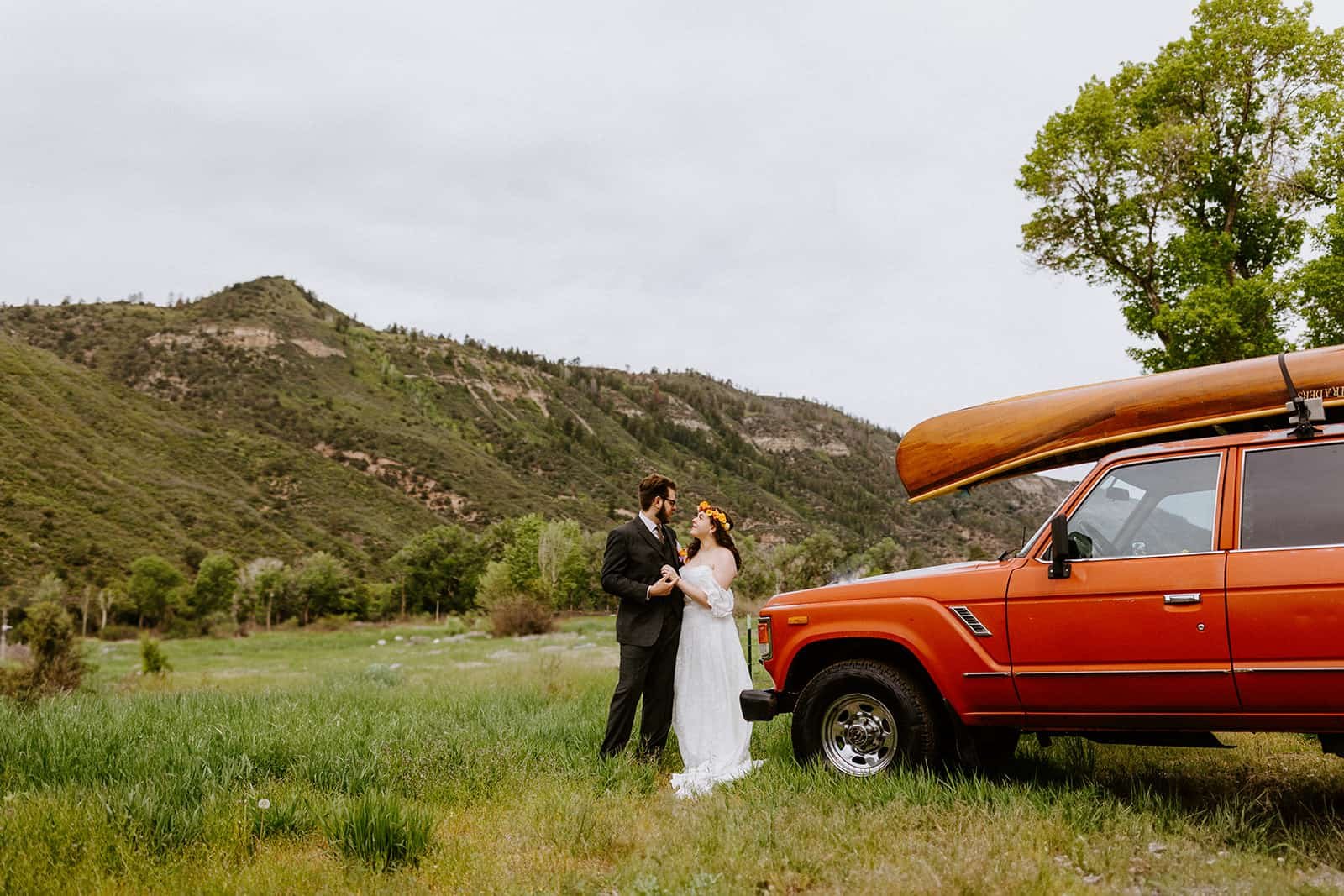 A man and woman in a wedding dress and suit smile and embrace in front of a red Toyota Land Cruiser