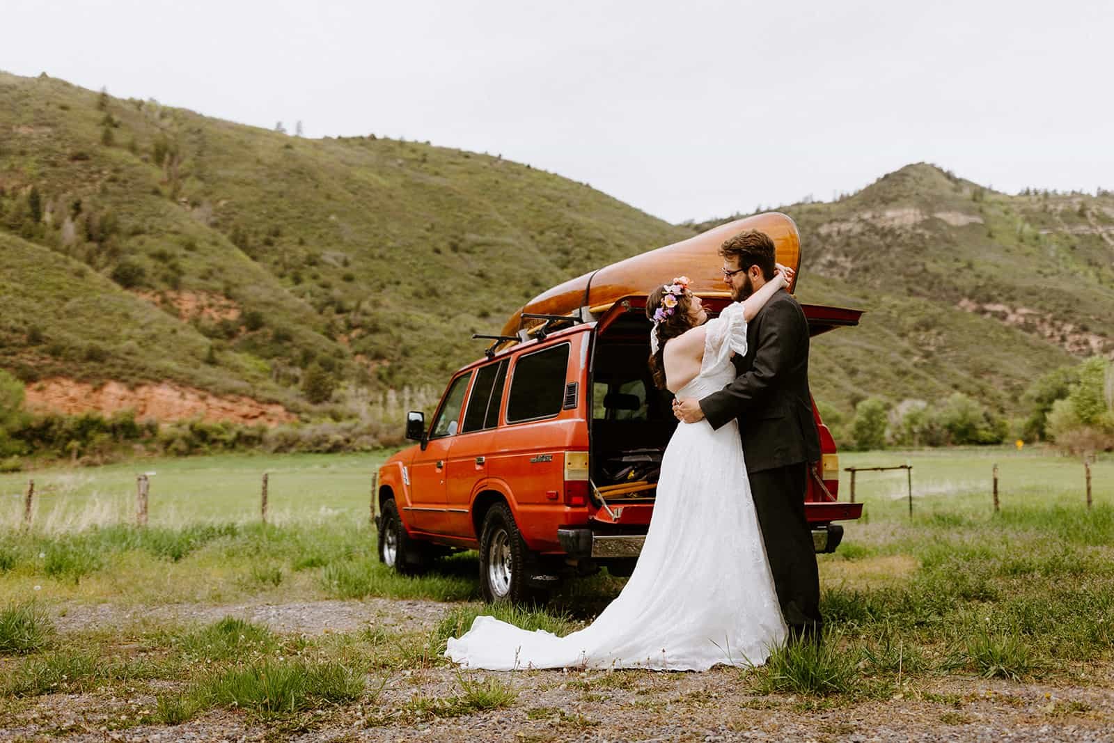 A man and woman in a wedding dress and suit smile and embrace in front of a red Toyota Land Cruiser