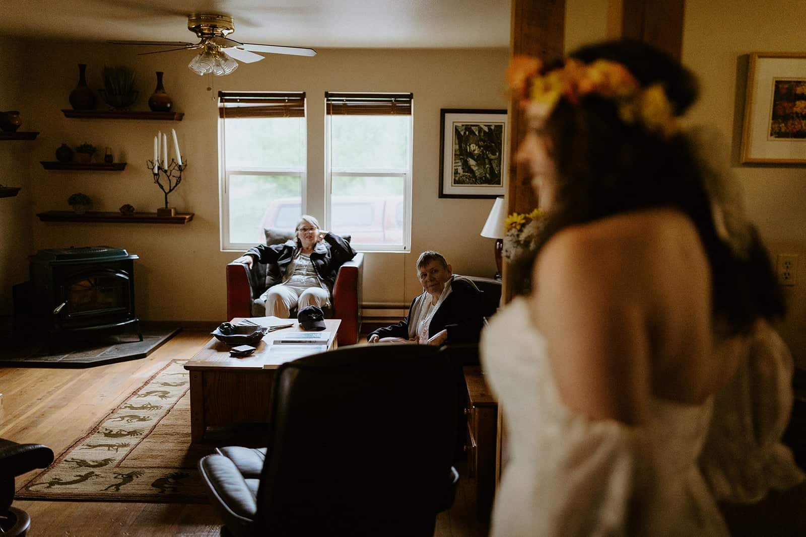 Two elderly women sitting in a living room look on as a woman walks in in her wedding dress