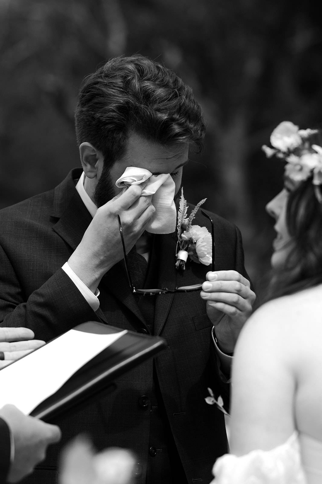 A man uses a tissue to wipe away a tear while he holds his glasses in his other hand at his wedding ceremony