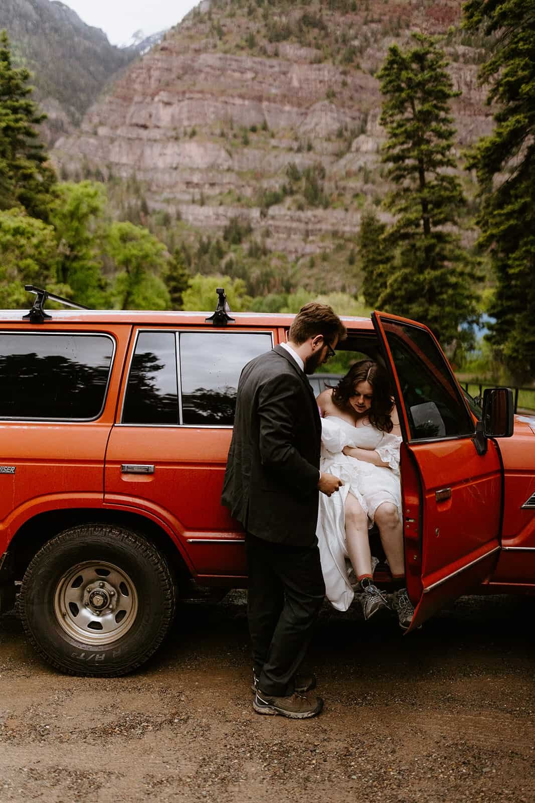 A man helps a woman in a wedding dress out of a red SUV