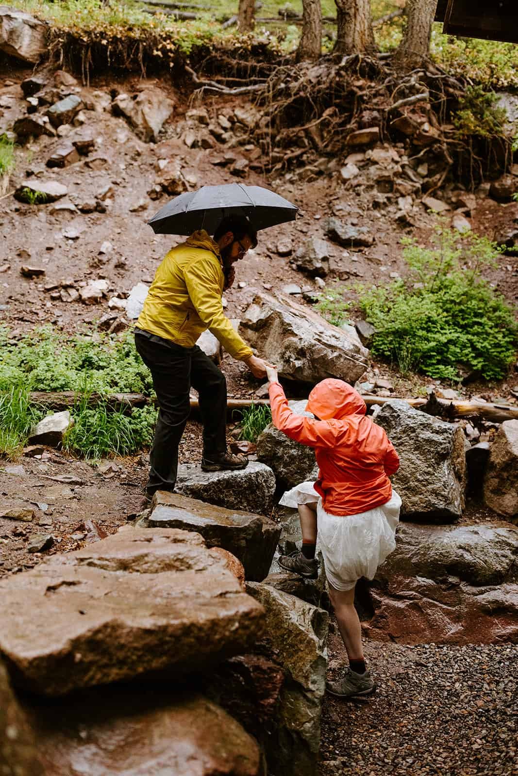 A man holding an umbrella helps a woman in a wedding dress and rain jacket up a rock ledge