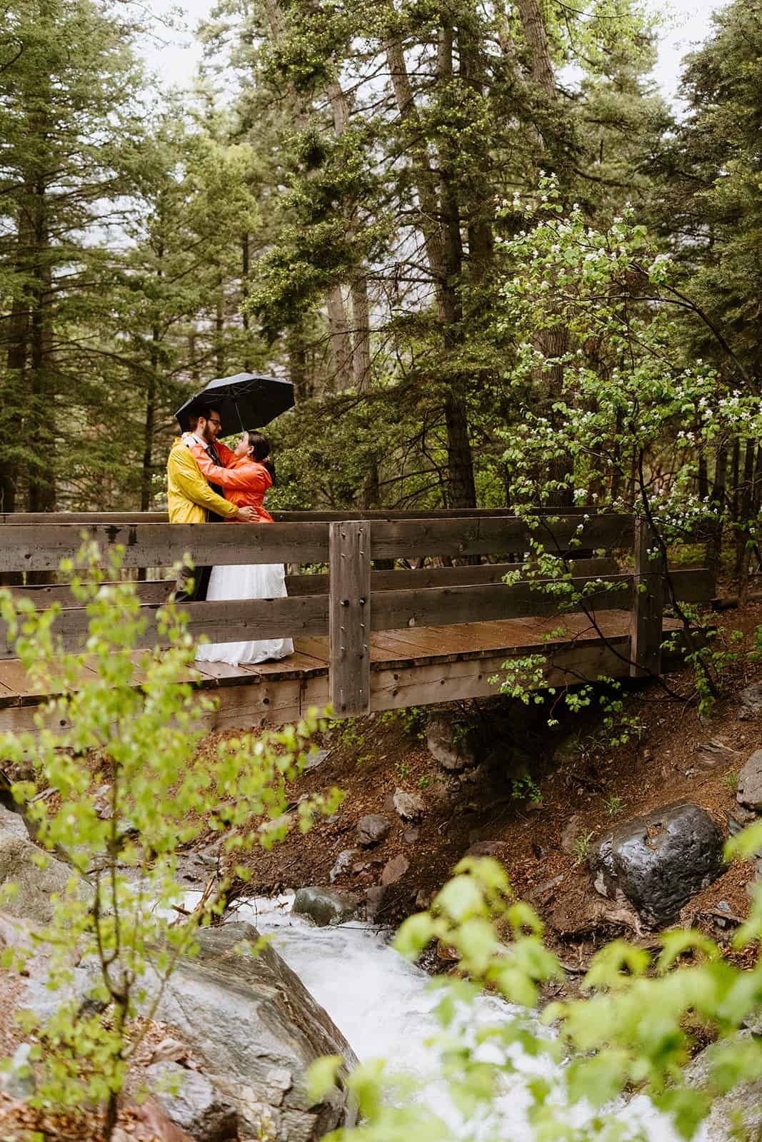 A couple in wedding attire stand on a bridge under an umbrella over a creek smiling at each other