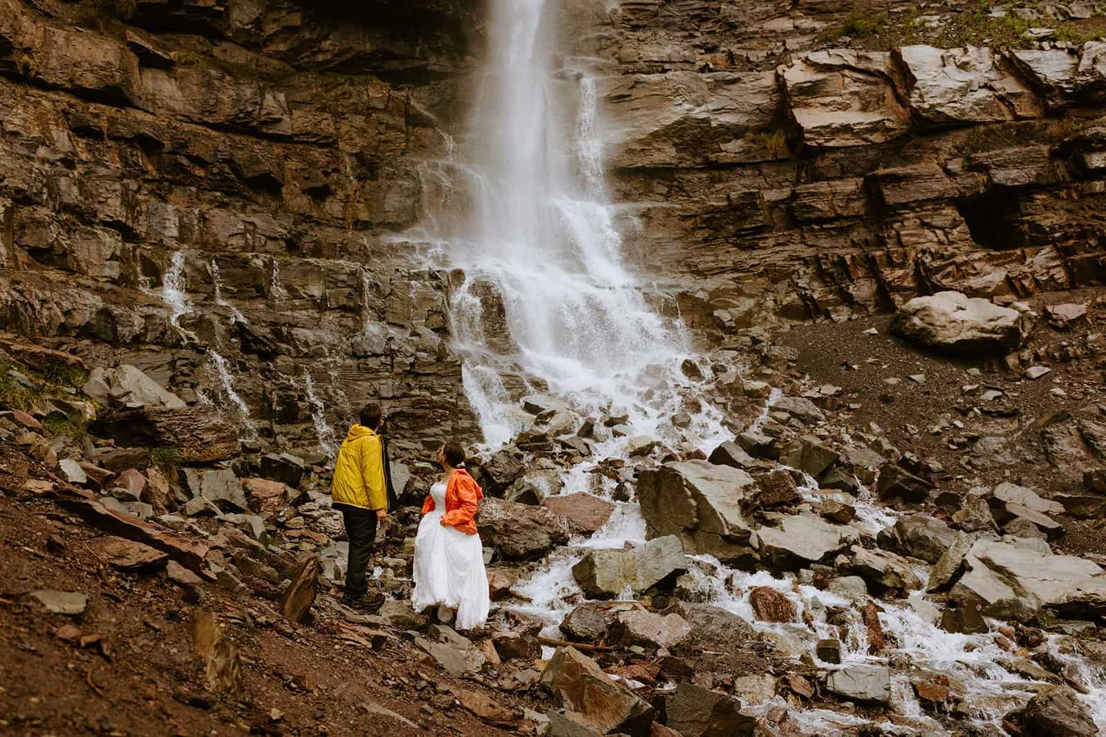 A couple in wedding clothes stands underneath a Colorado waterfall
