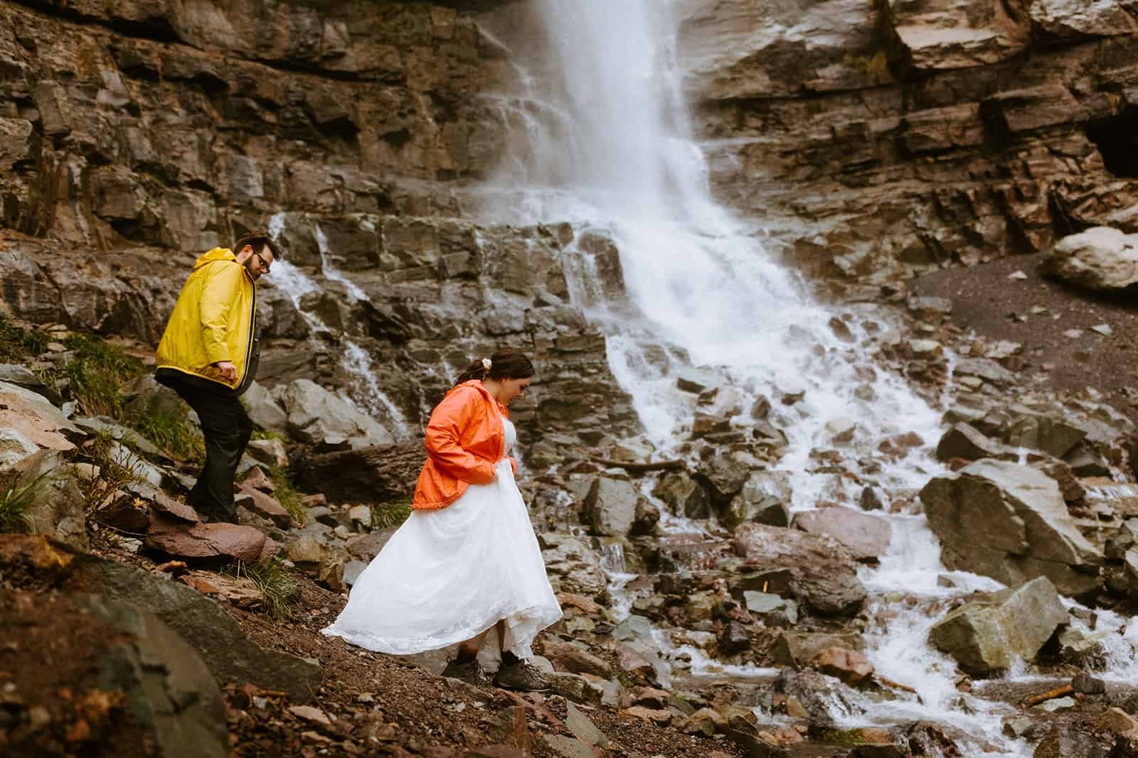 A woman holds up her wedding dress as she walks through the rocks underneath a Cascading waterfall in Ouray
