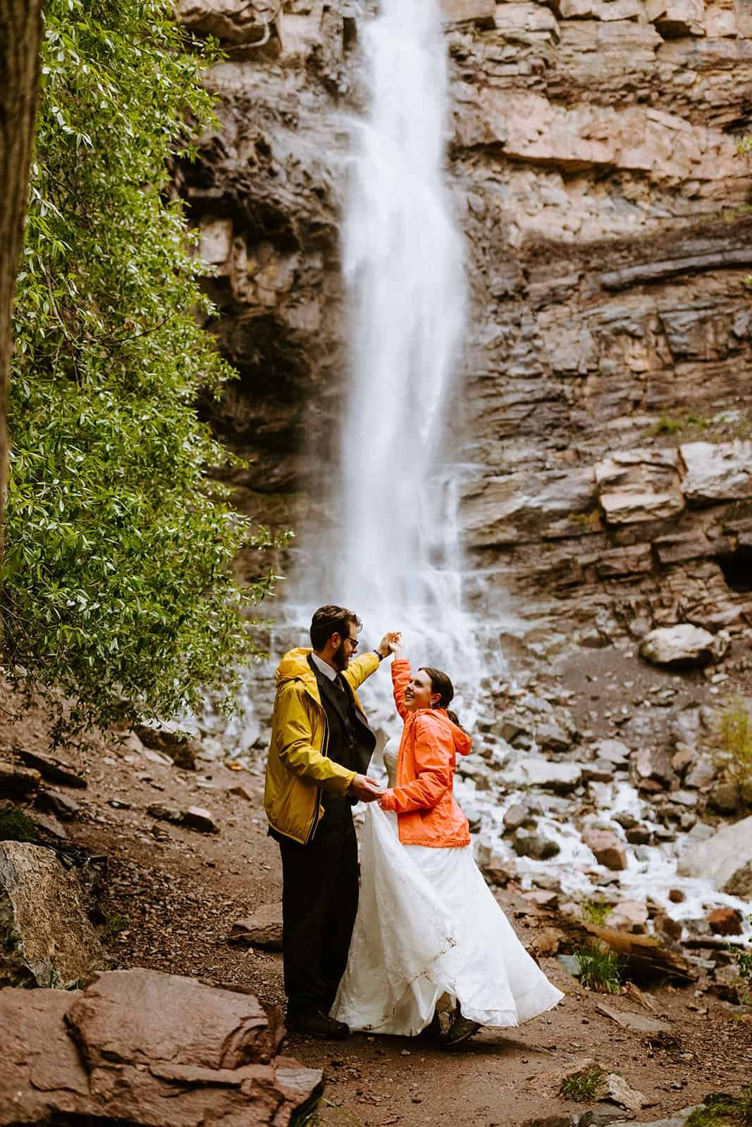 A couple in a wedding dress and suit dance in front of a waterfall in Ouray