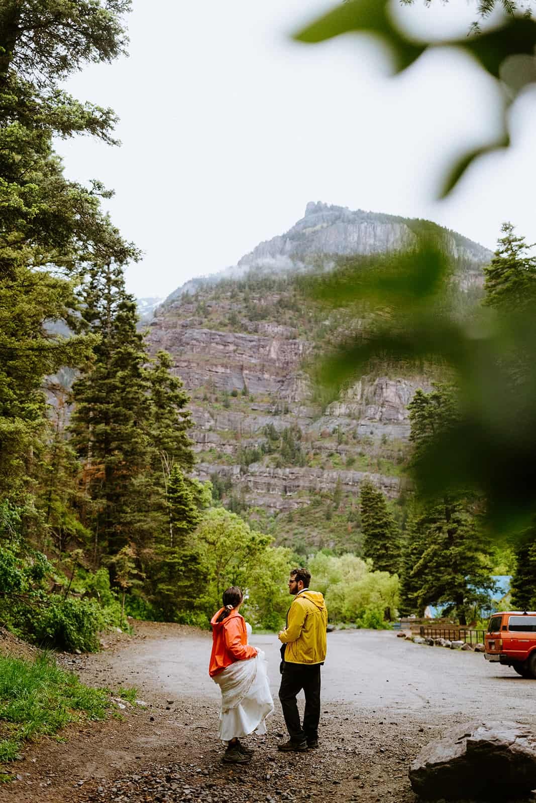 A man in a suit and woman in a wedding dress wear rain jackets and look at the town of Ouray from the Cascade Falls parking lot